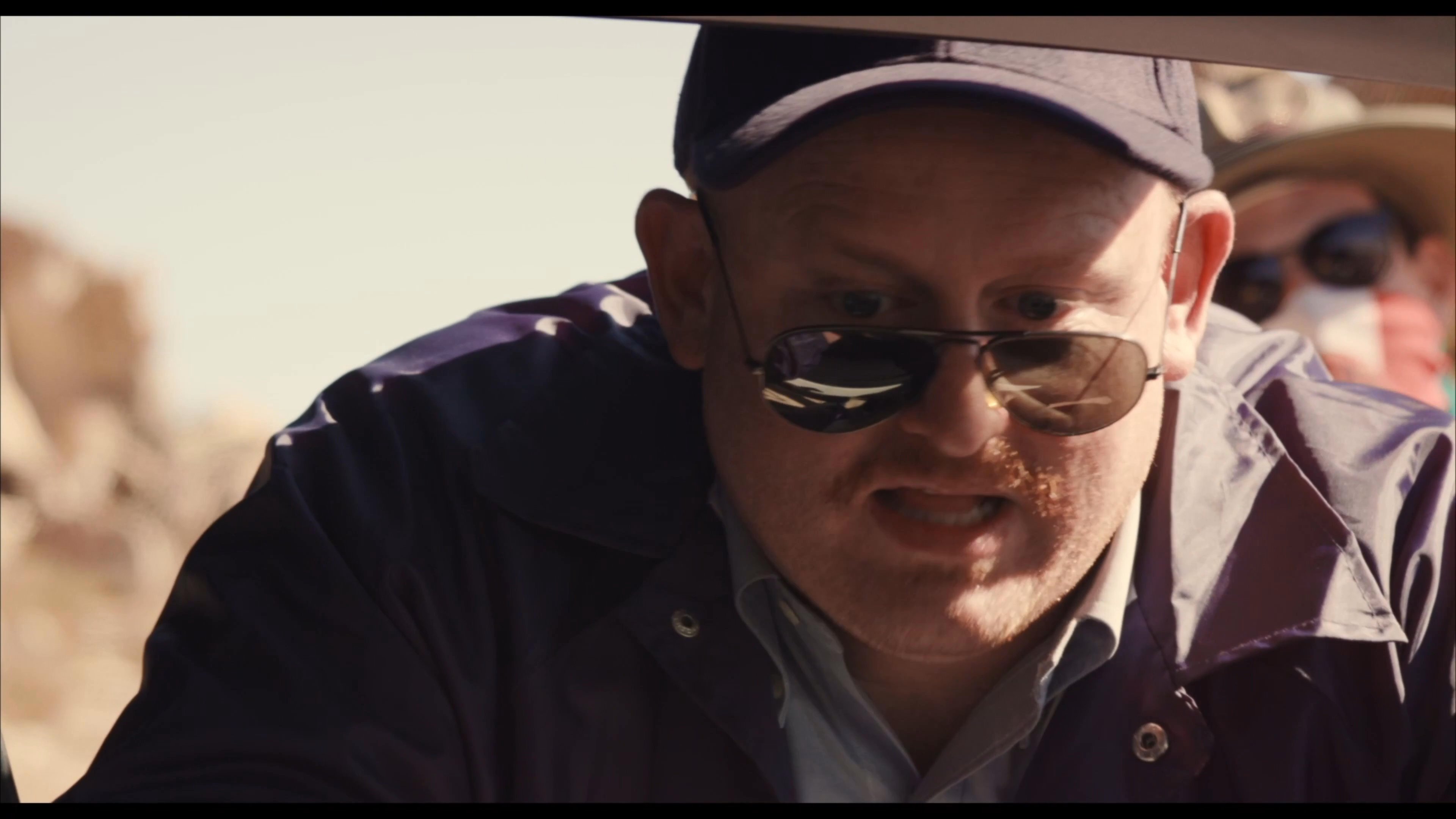 An agent wearing tactical gear and sunglasses conducts an intense field interrogation near a canine unit. The setting suggests an active investigation in a desert environment.