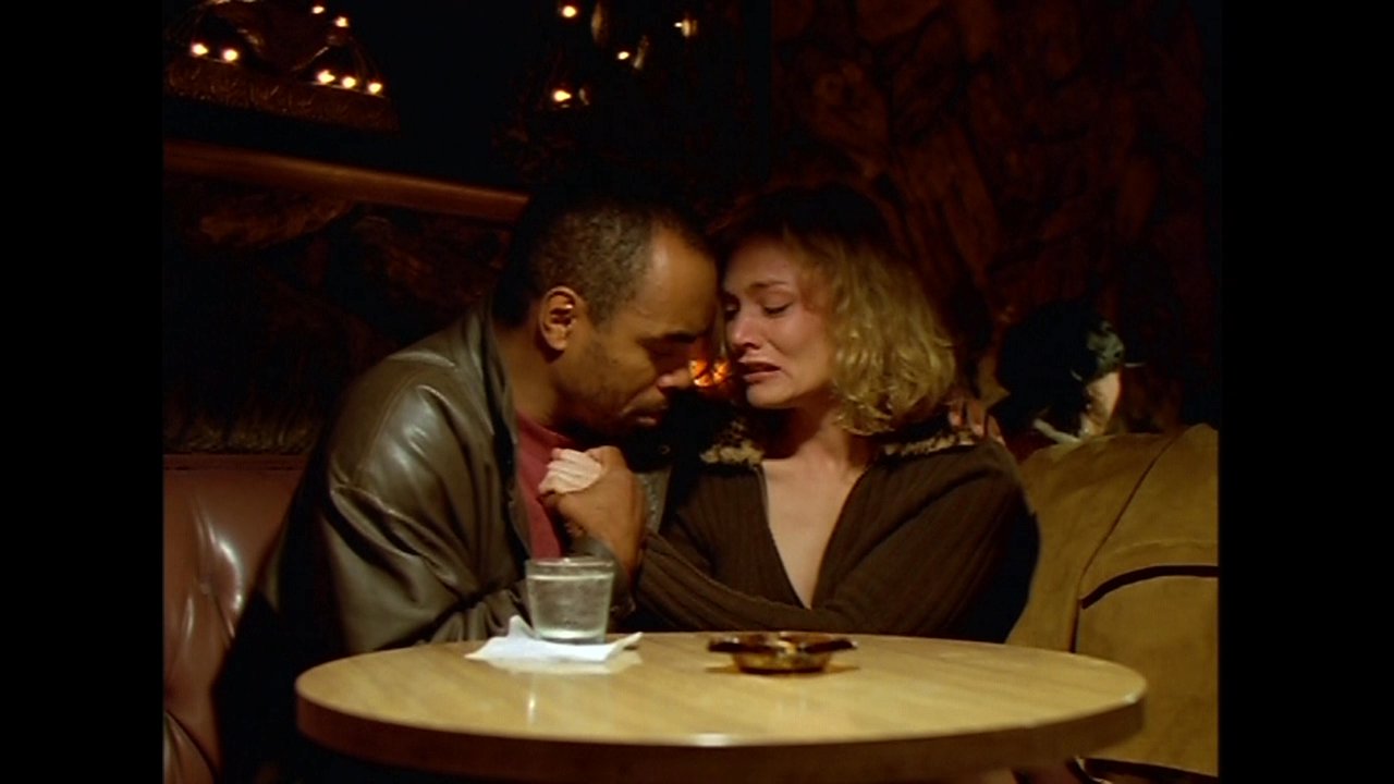 A man and a woman engage in an intimate, emotional conversation while seated together in a quiet, warmly lit diner booth.