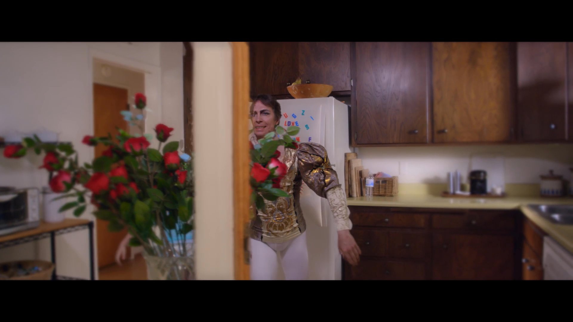 A father dressed in a ballet costume dances with his young daughter, who wears a princess dress, in their home. The sequence culminates in a playful, festive moment as red rose petals fall around the child.