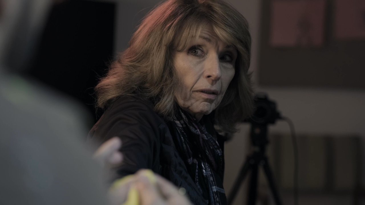 An artist engages in a focused discussion with a client within a dimly lit studio space equipped with easels and camera equipment. The dialogue centers on the creative process as they assess a portrait in progress.