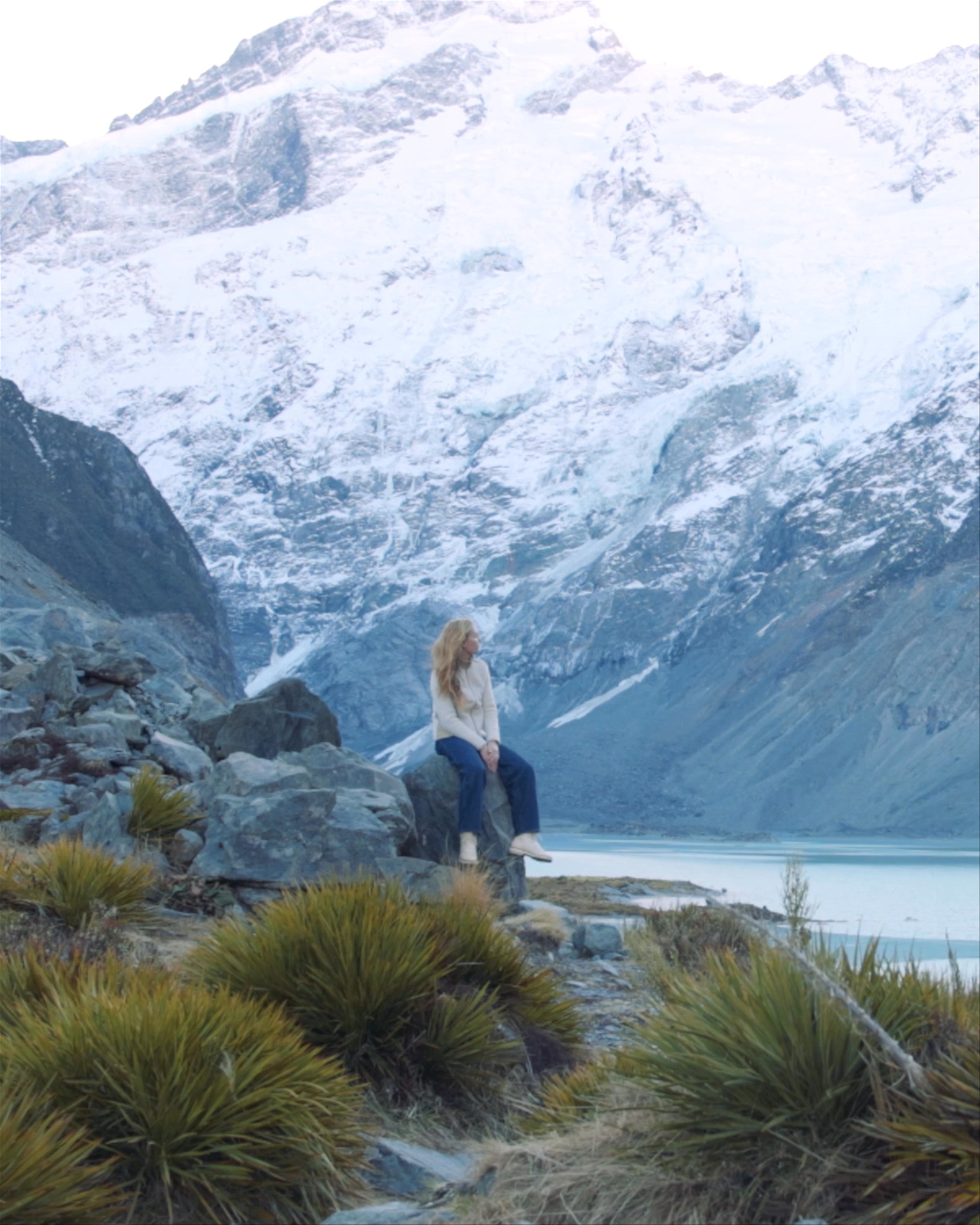 Woman sitting on rock alongside river and snowy mountains in New Zealand