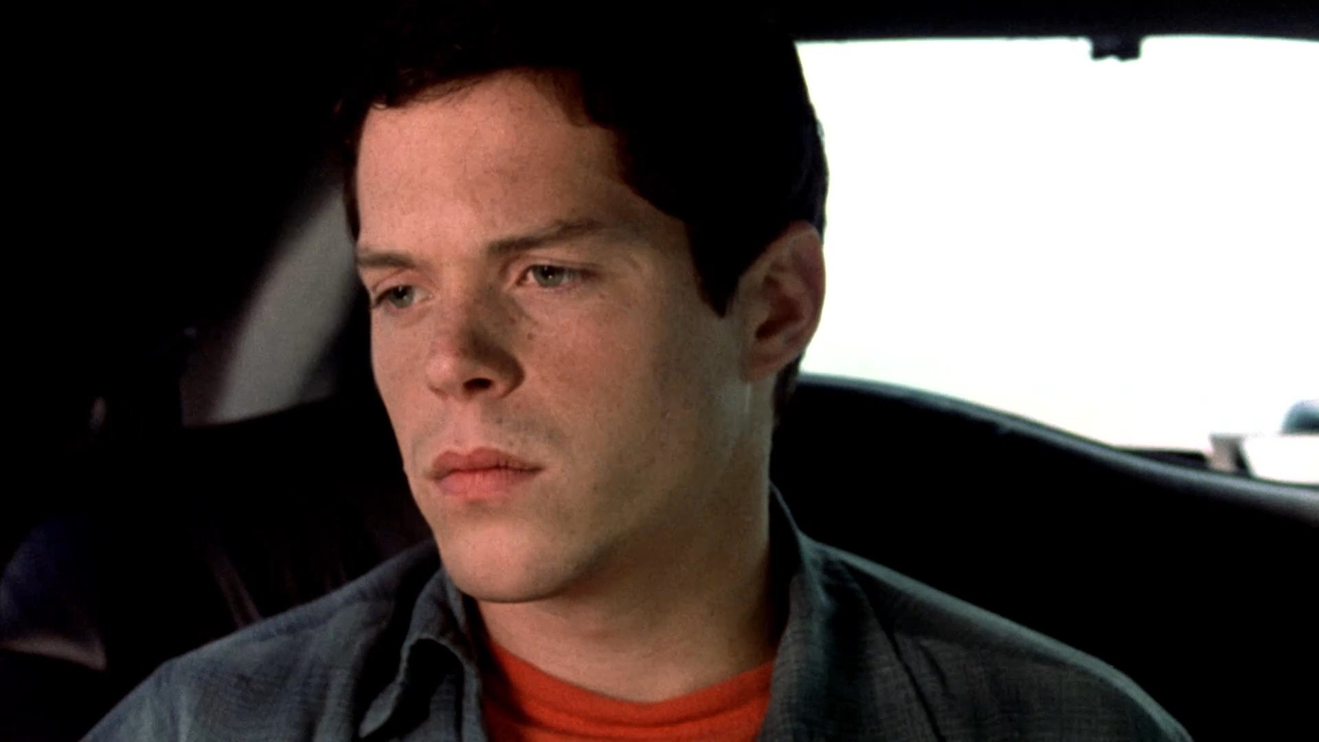 Two individuals engage in a somber conversation inside a limousine while watching a miniature diorama of a roadside accident on a small onboard television.
