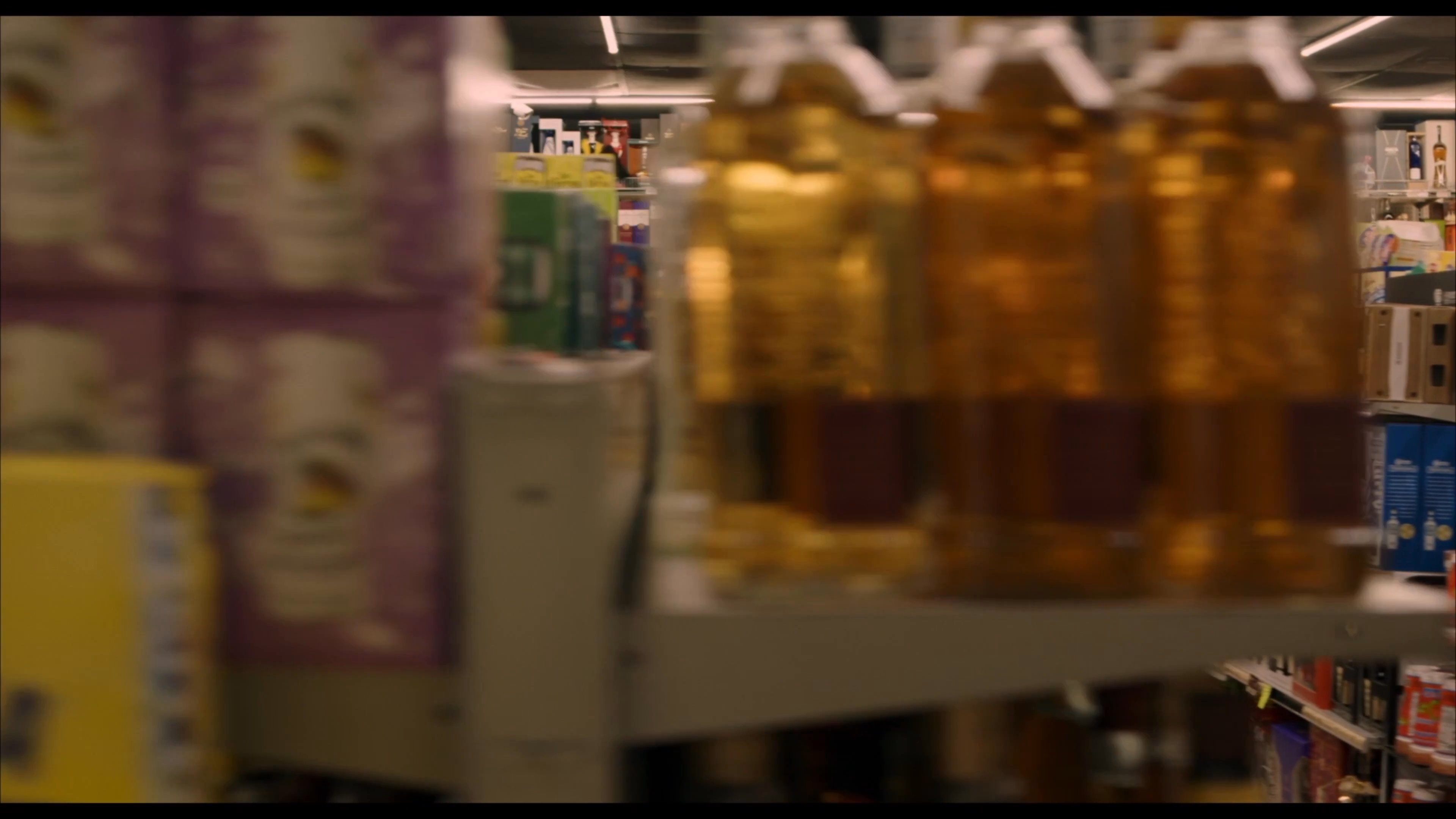 A woman reads a magazine behind the counter while a man walks through the aisles of a convenience store. The camera pans across the inventory of merchandise lining the store shelves.