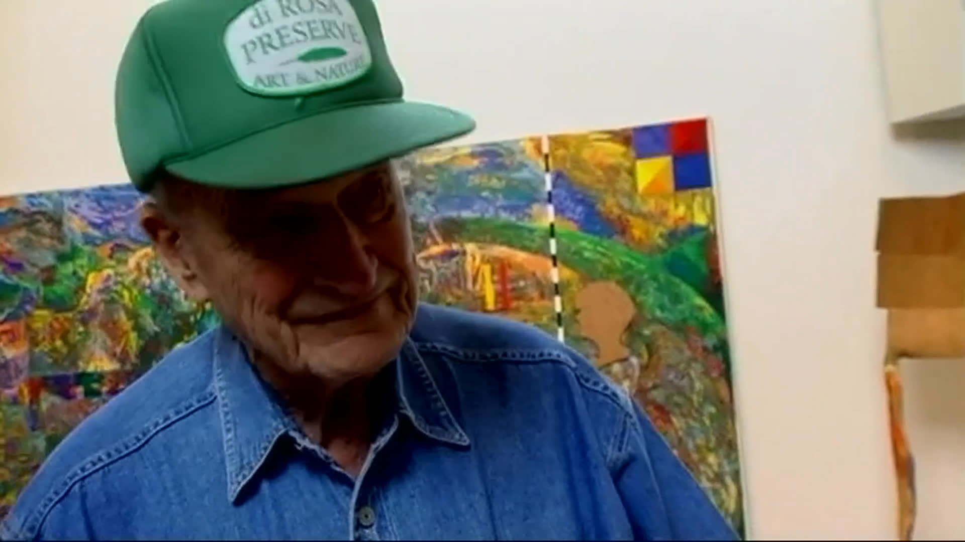 An elderly man in a green cap discusses his art collection among colorful paintings and sculptures, expressing a wish to share it nationwide while welcoming visitors to the gallery. He gestures toward displayed works as others handle a sculpture nearby.