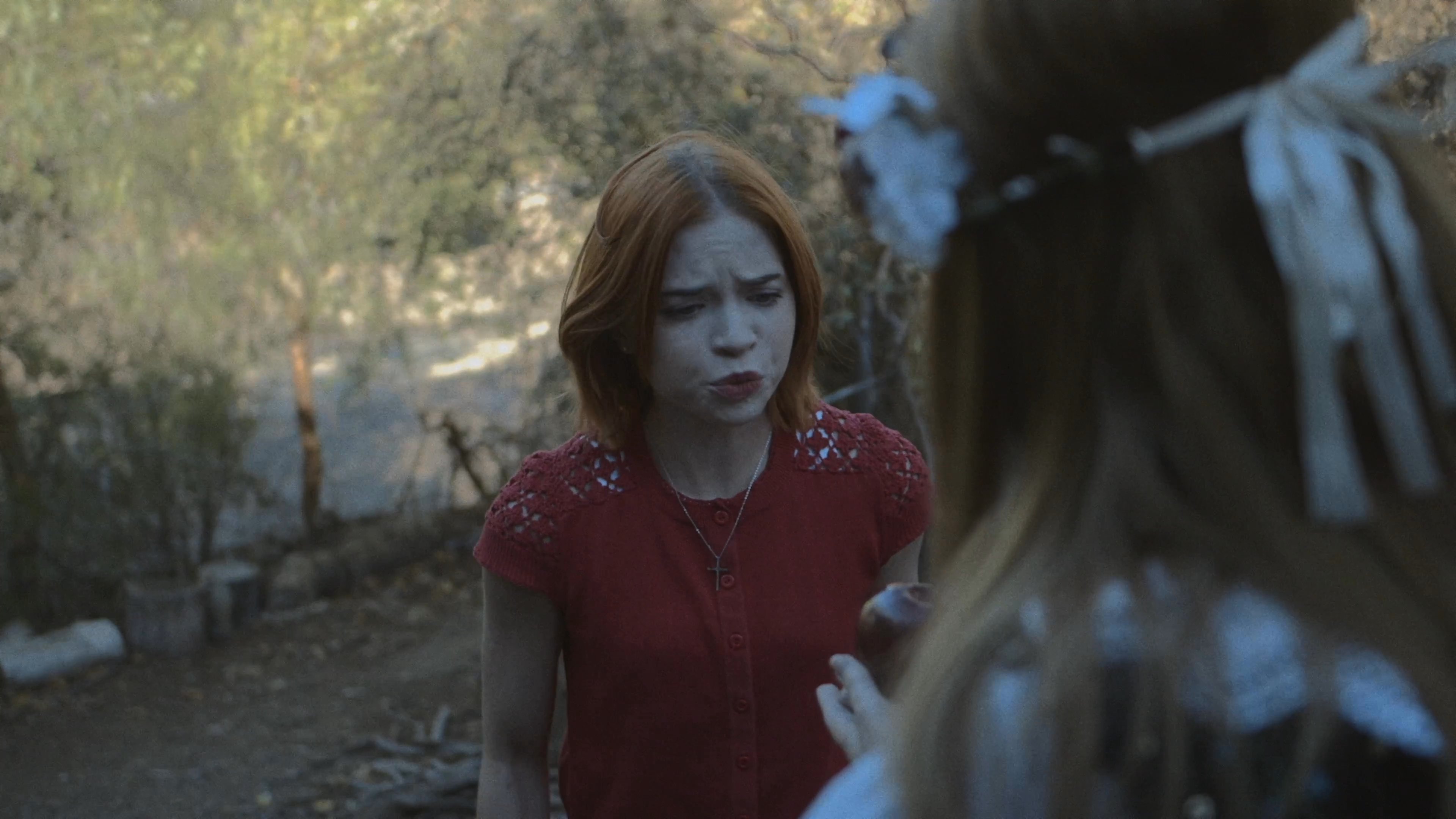 Two women, one dressed in a rustic floral headpiece and apron, present a red apple to another woman within a dense, wooded environment. Their interaction evokes a classic folklore narrative characterized by mystery and dramatic tension.