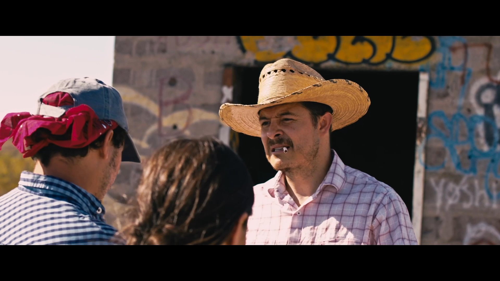 A man in a cowboy hat aggressively confronts a couple in a remote, graffiti-marked desert location regarding a sum of money.