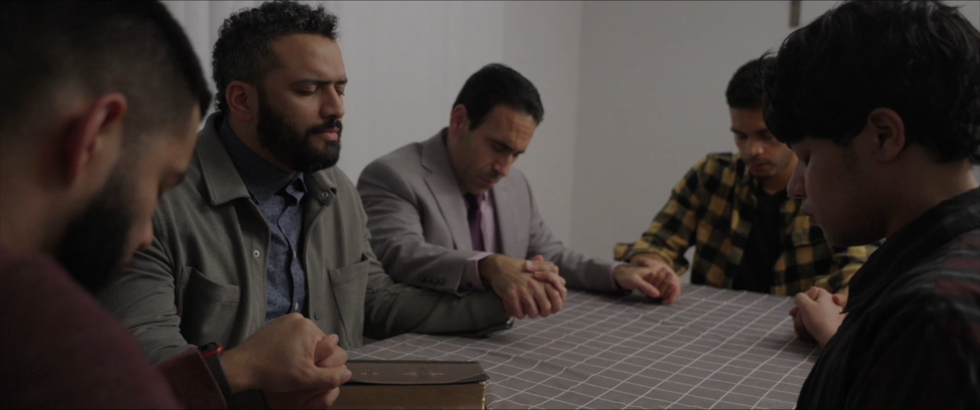 A diverse group of men sits around a table to pray and engage in a collective spiritual reflection. The individuals join hands in a quiet, contemplative atmosphere.