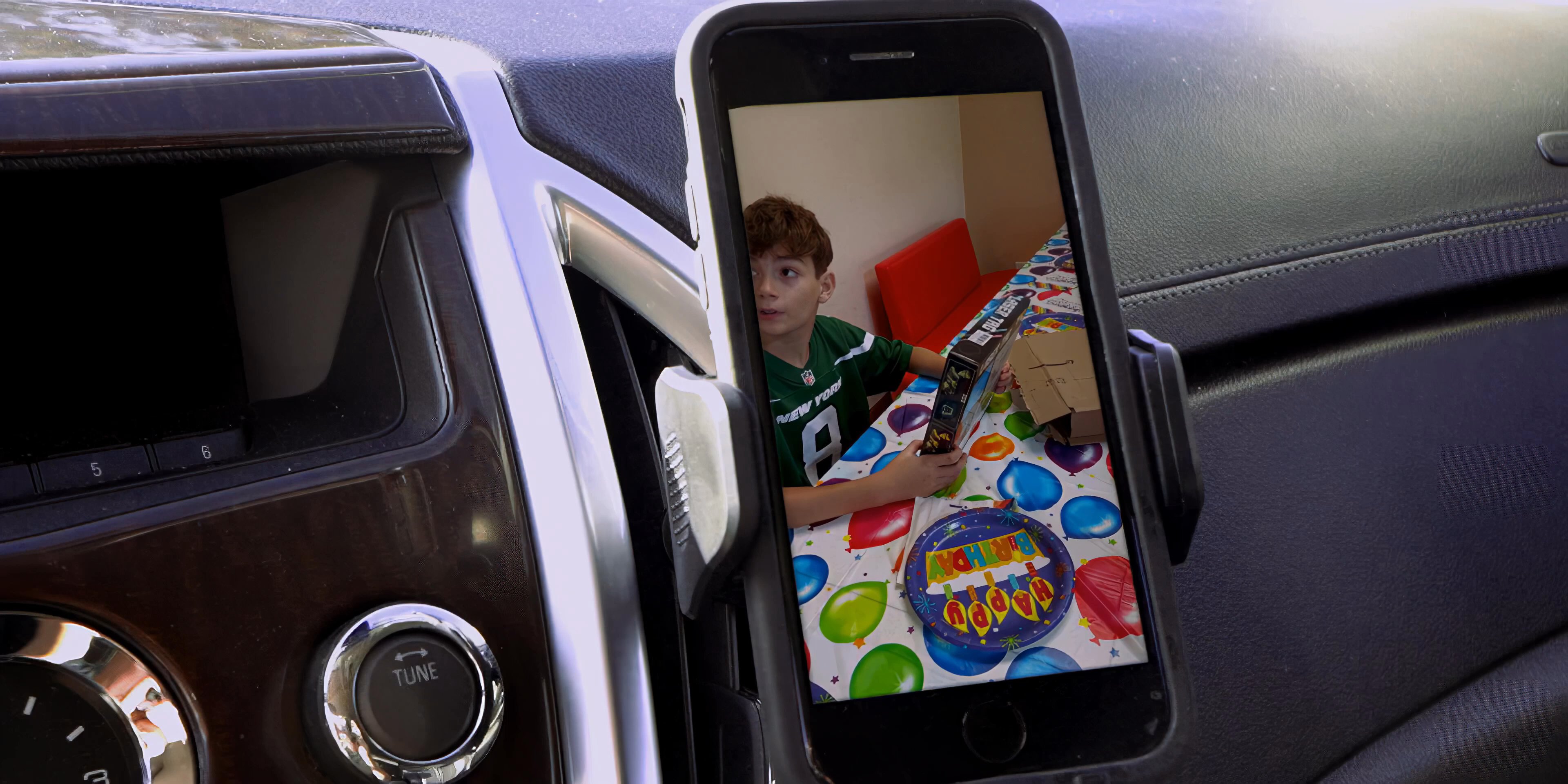 A young child excitedly opens a birthday gift while the scene is captured and displayed on a smartphone screen mounted in a vehicle. A man driving the car observes the digital transmission during his commute.
