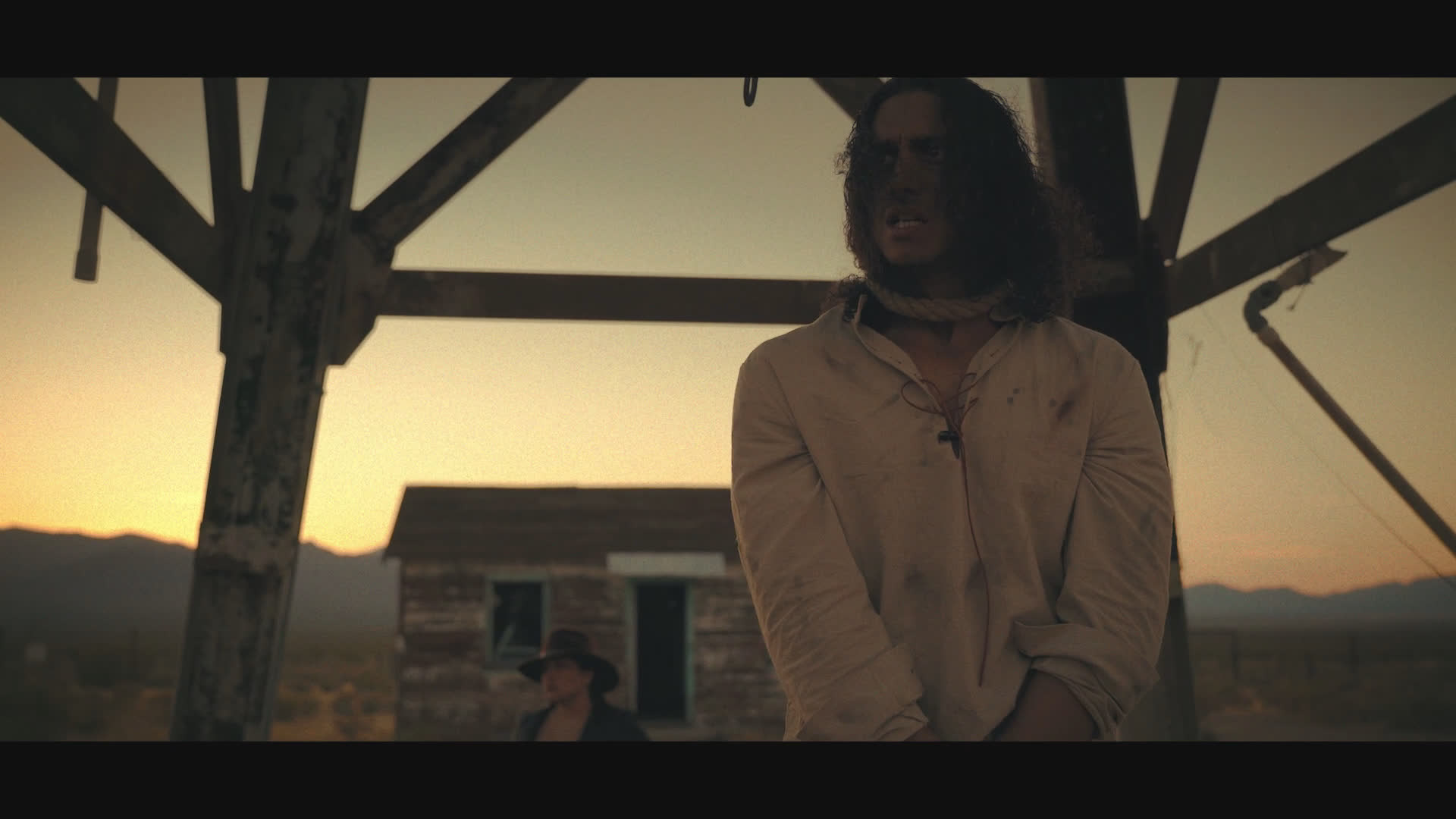 A long-haired man stands beneath a weathered wooden shelter as a tense exchange unfolds against a dusty settlement at sunset. He reacts with disbelief to questions and an accusation about waiting to rob him.