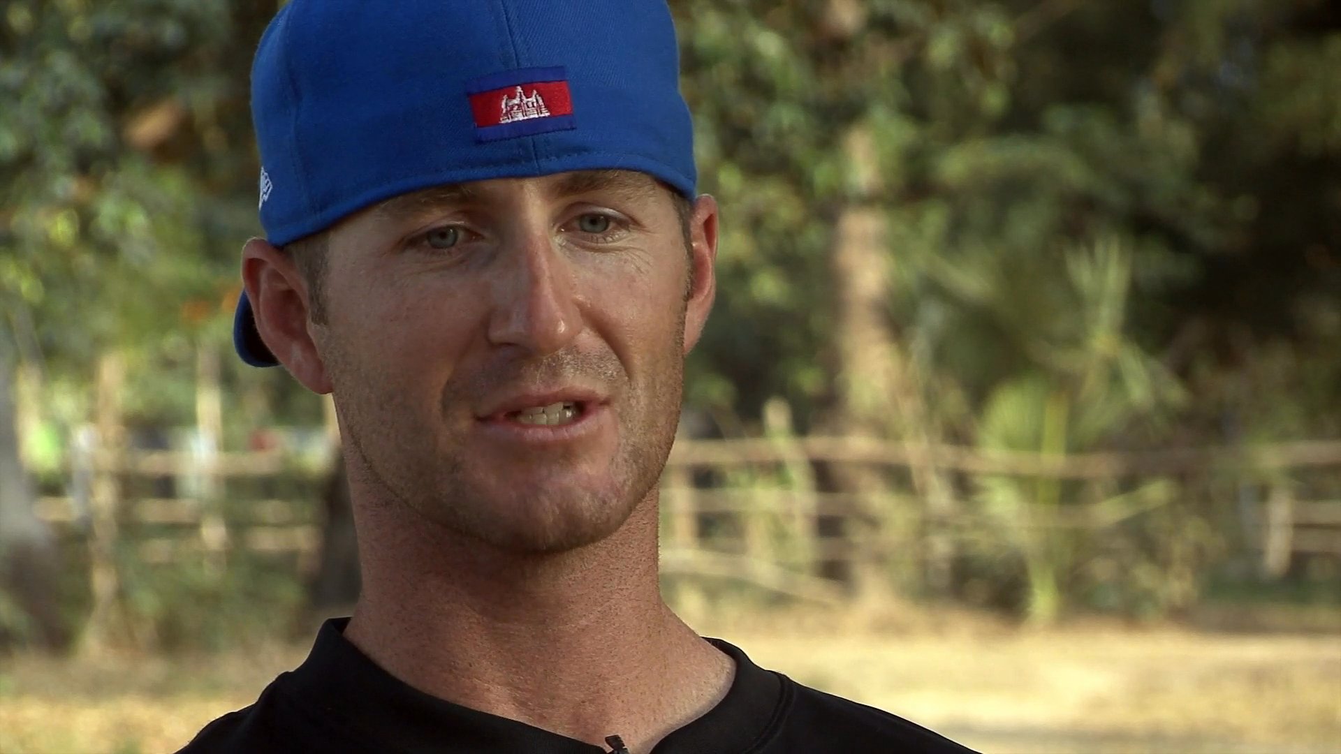 A man wearing a blue baseball cap speaks directly to the camera in an outdoor setting.