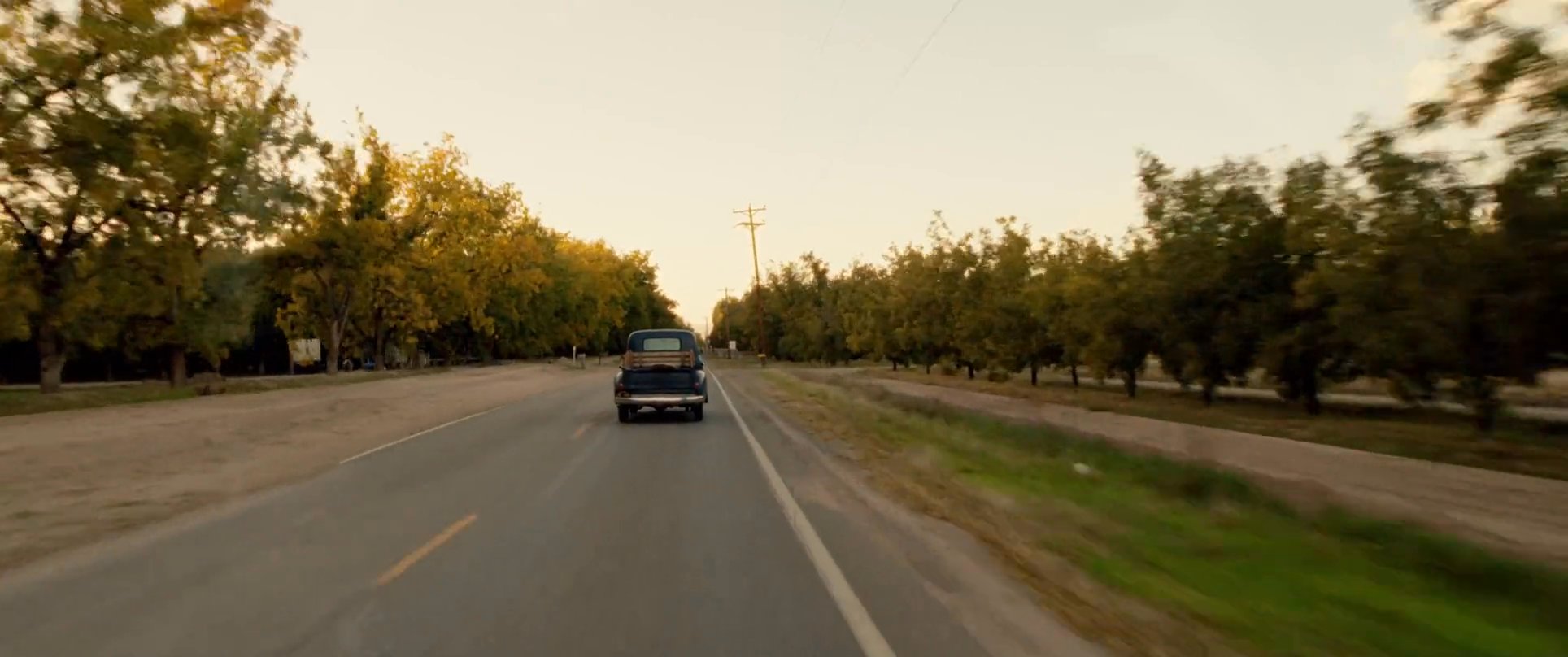 A man drives a vintage truck through the scenic landscapes of Mesilla, New Mexico, passing historic structures and lush orchards during golden hour.