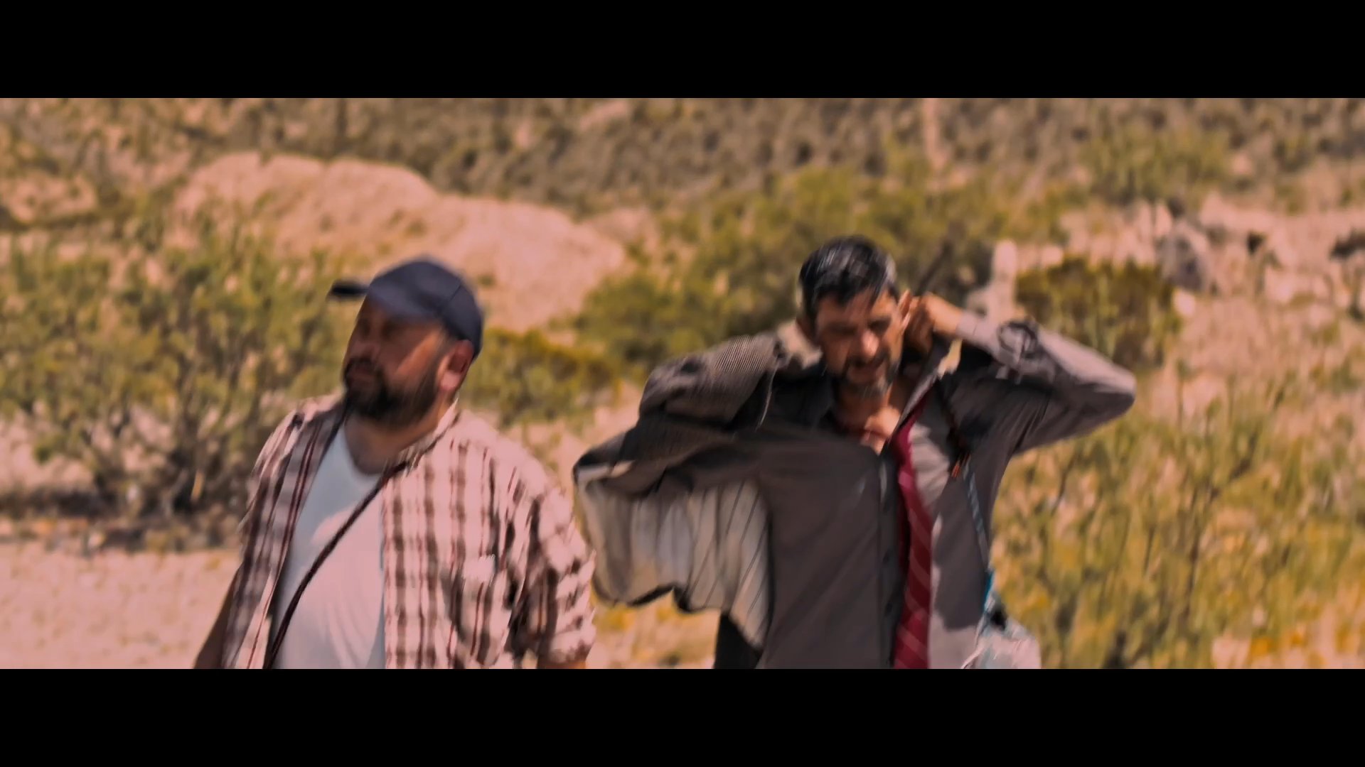Two men navigate a challenging, arid desert environment under intense heat. They continue their journey across the rugged, rocky terrain, exhibiting signs of physical exertion.