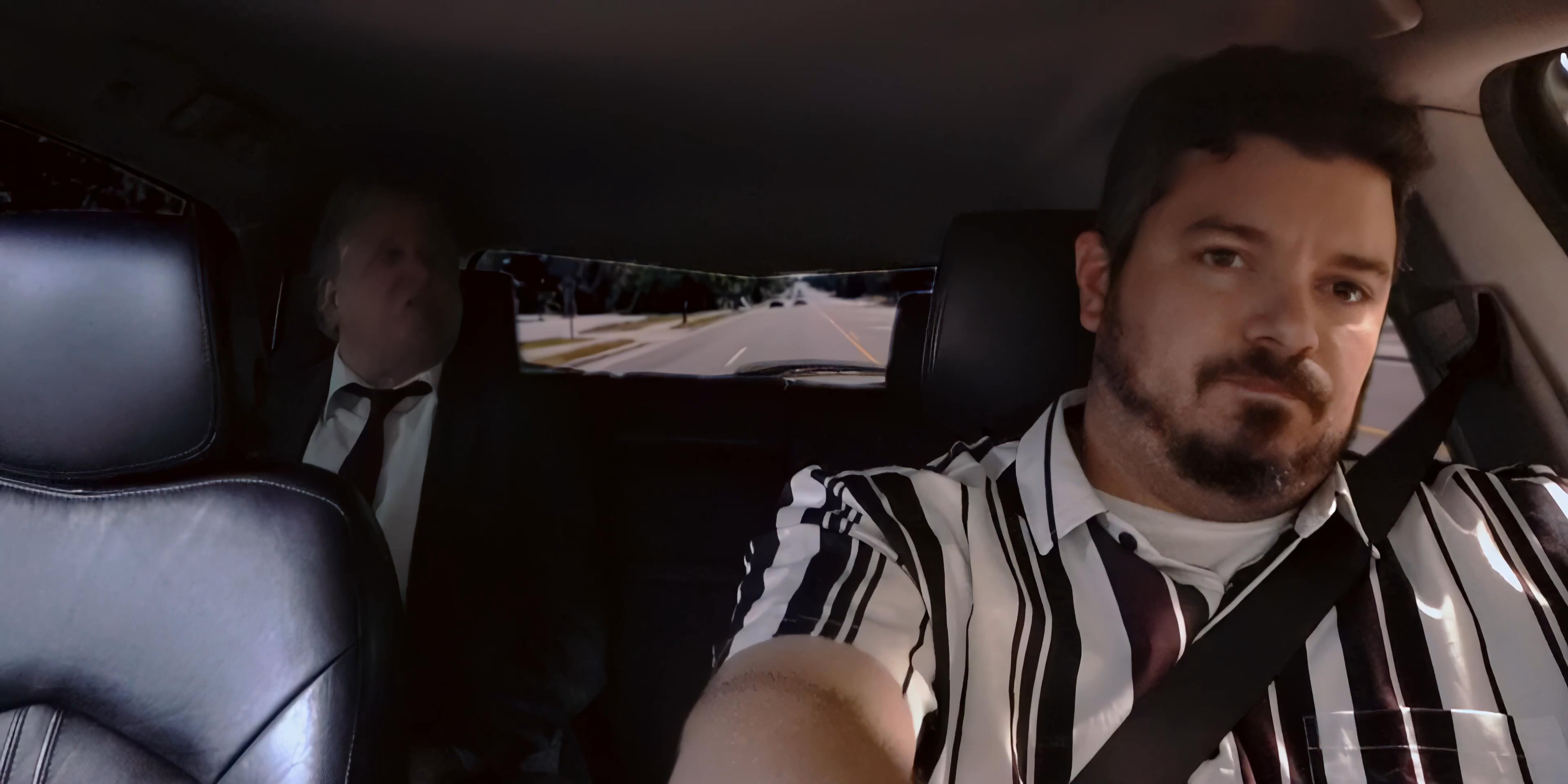 Two men travel in a car, with one acting as a passenger and the other as the driver, while engaged in a conversation during a daytime commute.