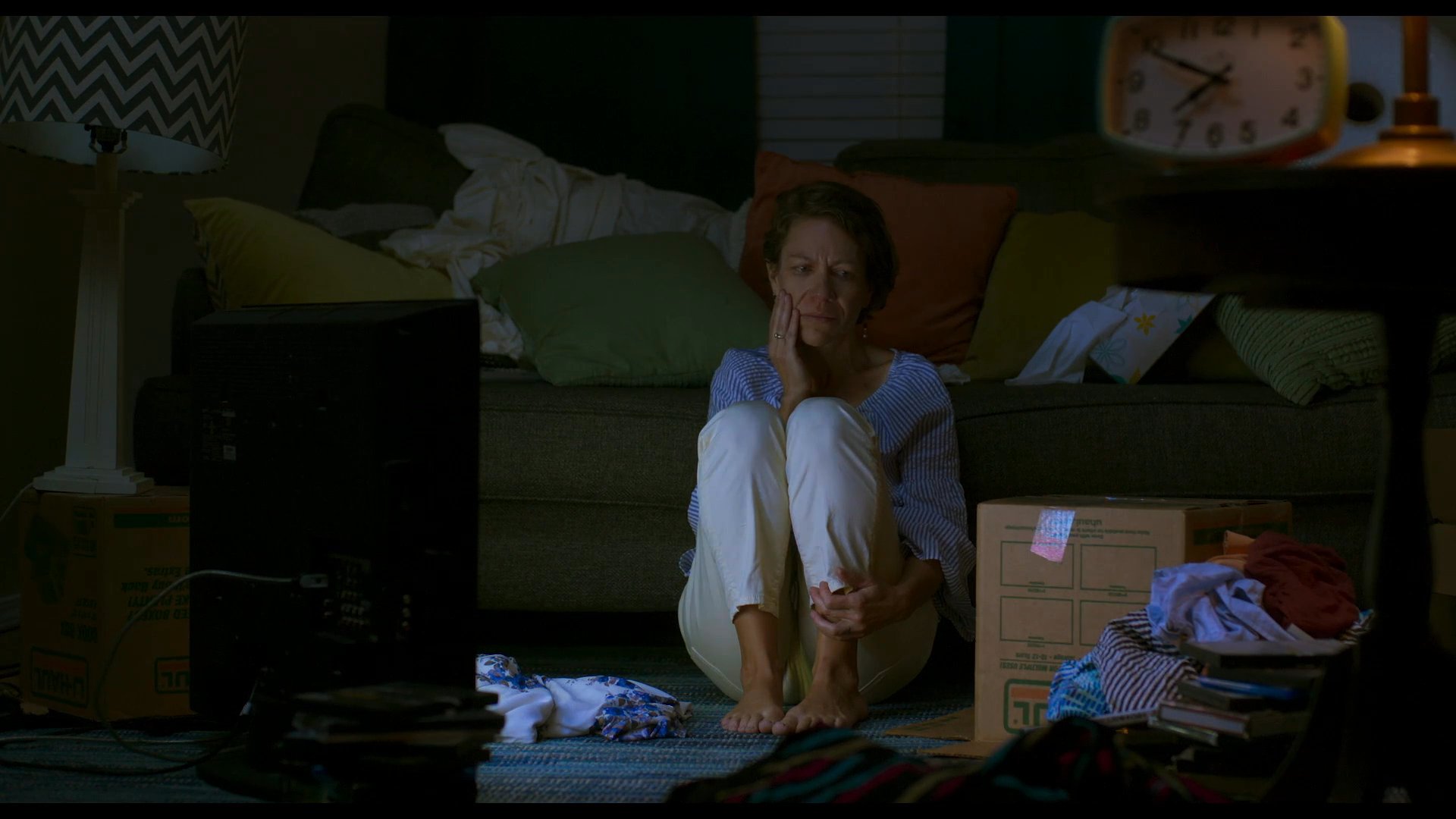 A woman sits quietly on the floor of a dimly lit living room, lost in deep thought amidst boxes and personal belongings. The scene conveys a sense of stillness and personal reflection within a domestic space.