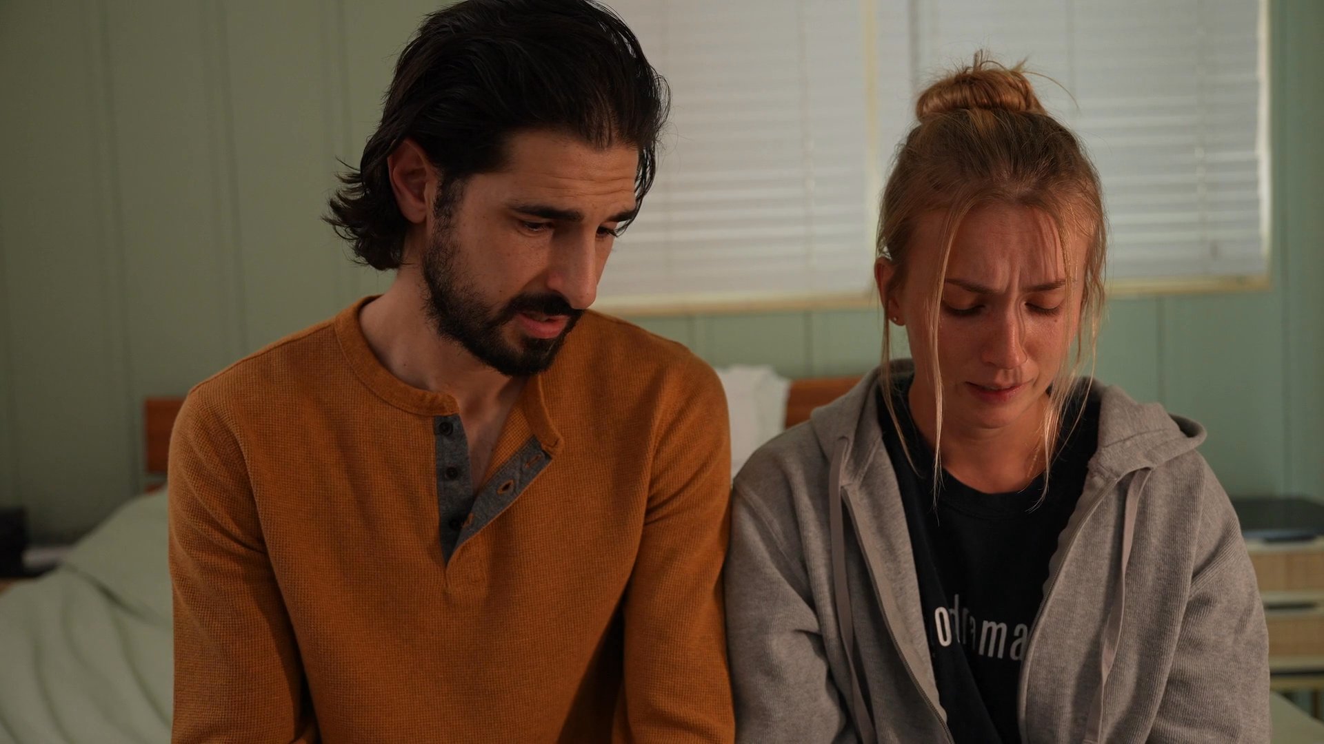A man in an orange shirt sits on a bed and provides emotional support to a crying woman as they engage in a serious, private conversation.