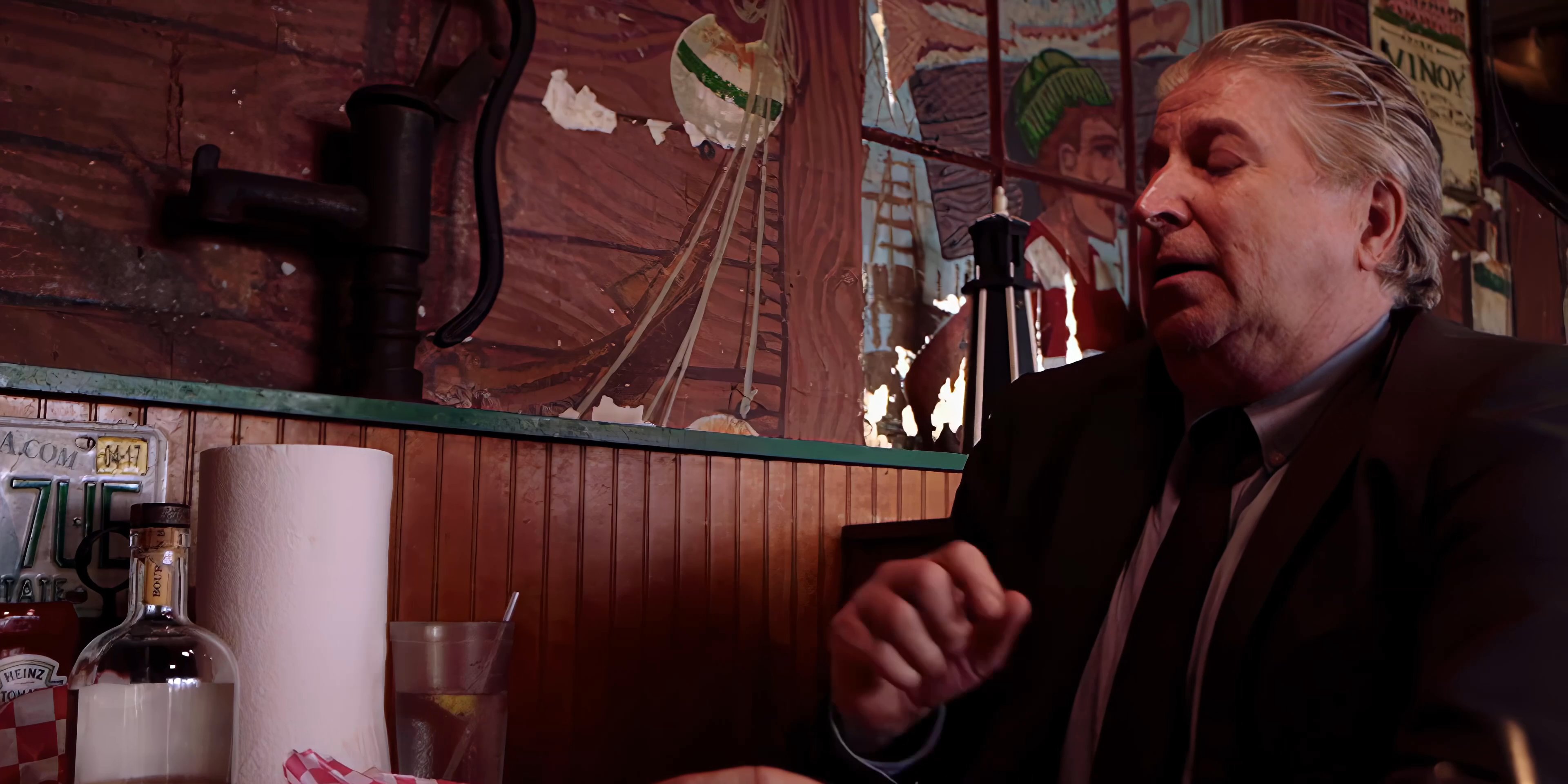 Two men engage in conversation inside a vintage-style diner while one pours a drink from a glass bottle.