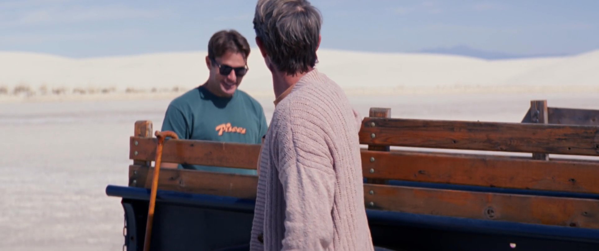 Two men relax by a pickup truck in a vast white sand desert, sharing a drink and preparing to unload their surfboard.
