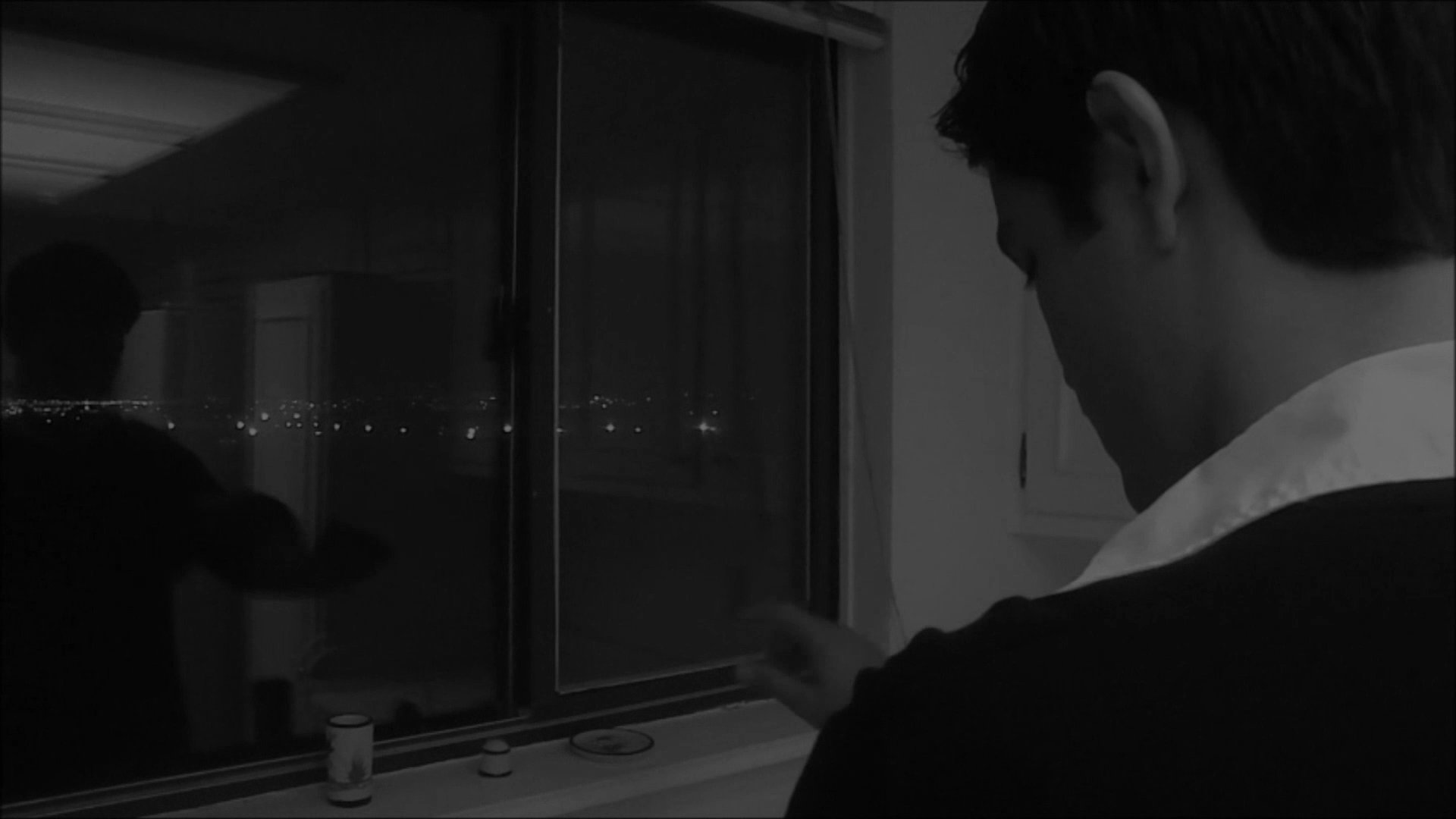 A man moves through a quiet kitchen at night, eventually pausing in deep contemplation while gazing out of a darkened window.