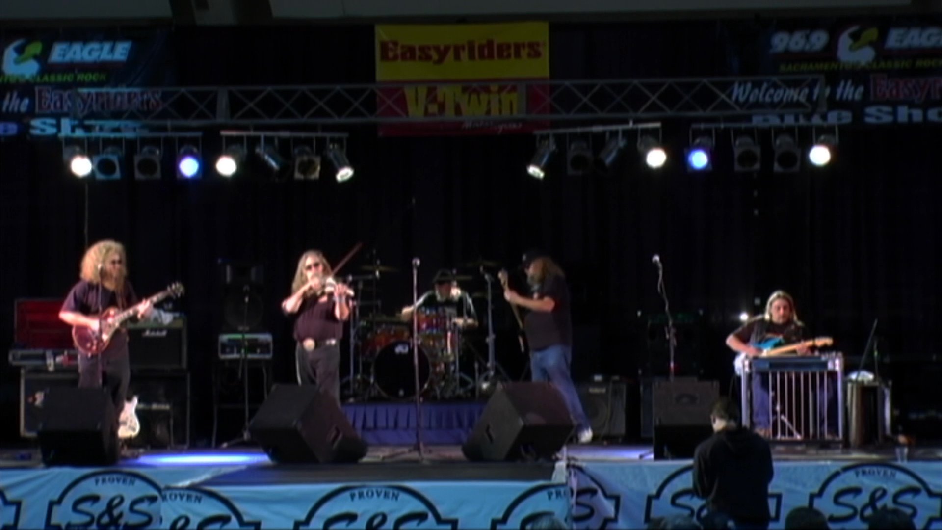 A long-haired violinist plays an electric instrument during a stage performance with a musical ensemble. The sequence shifts between close-up shots of the musician and wide angles showing the full band in a concert setting.