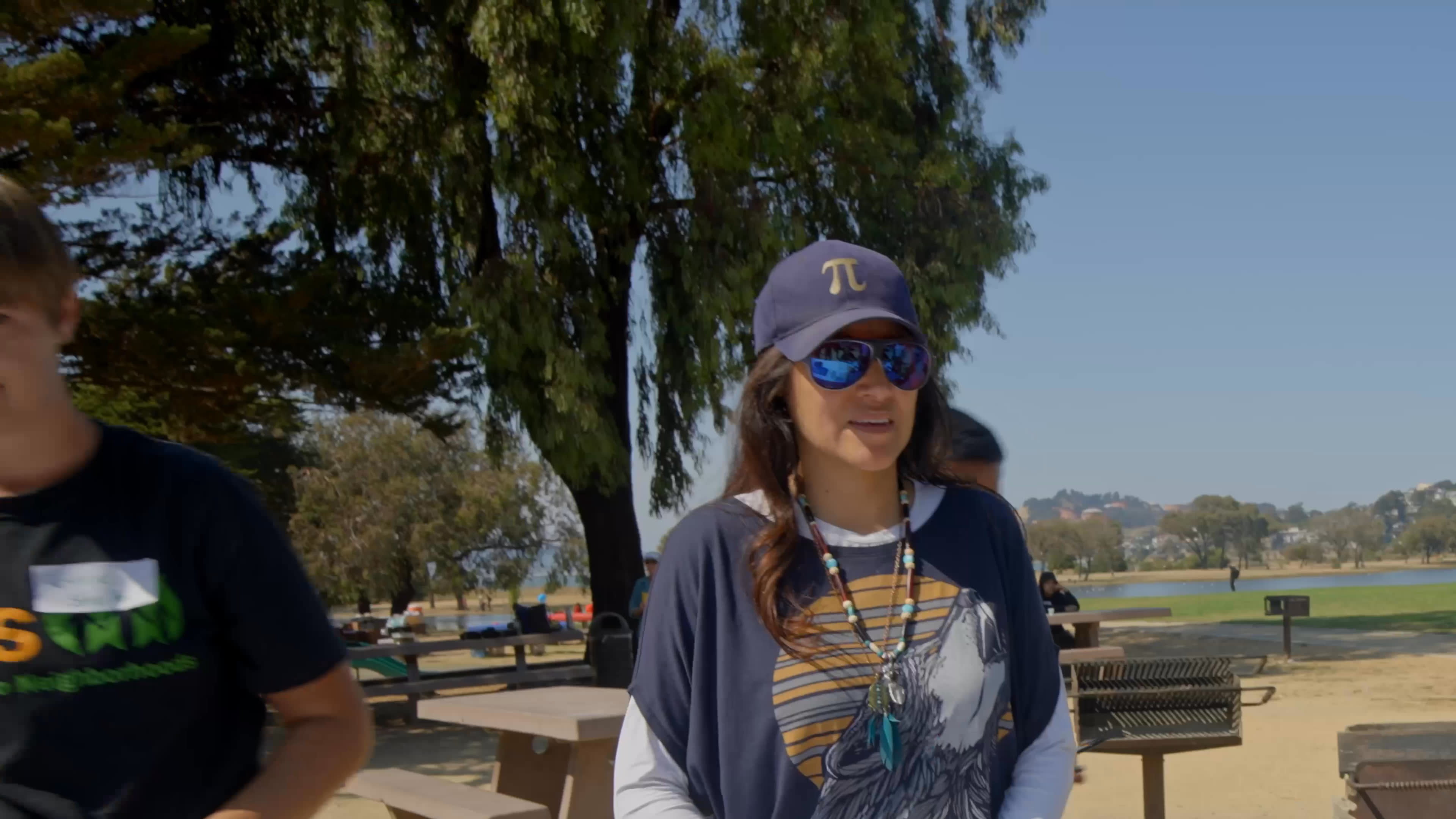 At a sunny park gathering, a woman in a cap and sunglasses joins a group activity, then speaks about being born in Mexico City and the challenges immigrants face. Participants gather around a picnic table as the conversation continues.