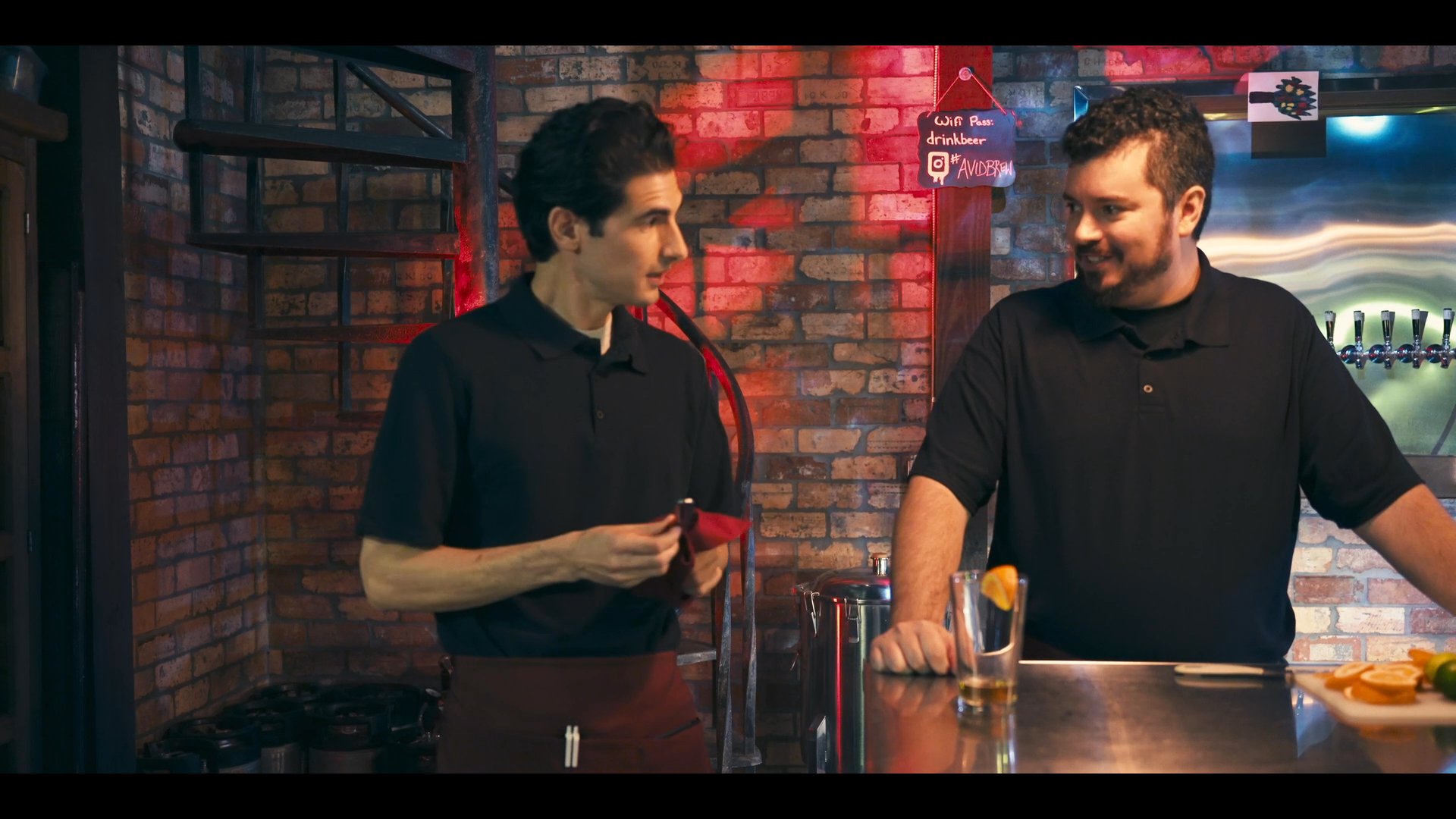 Two bartenders coordinate tasks behind a bar counter, discussing drink preparation before one moves to a different area of the establishment. The professional setting features brick decor and warm accent lighting.