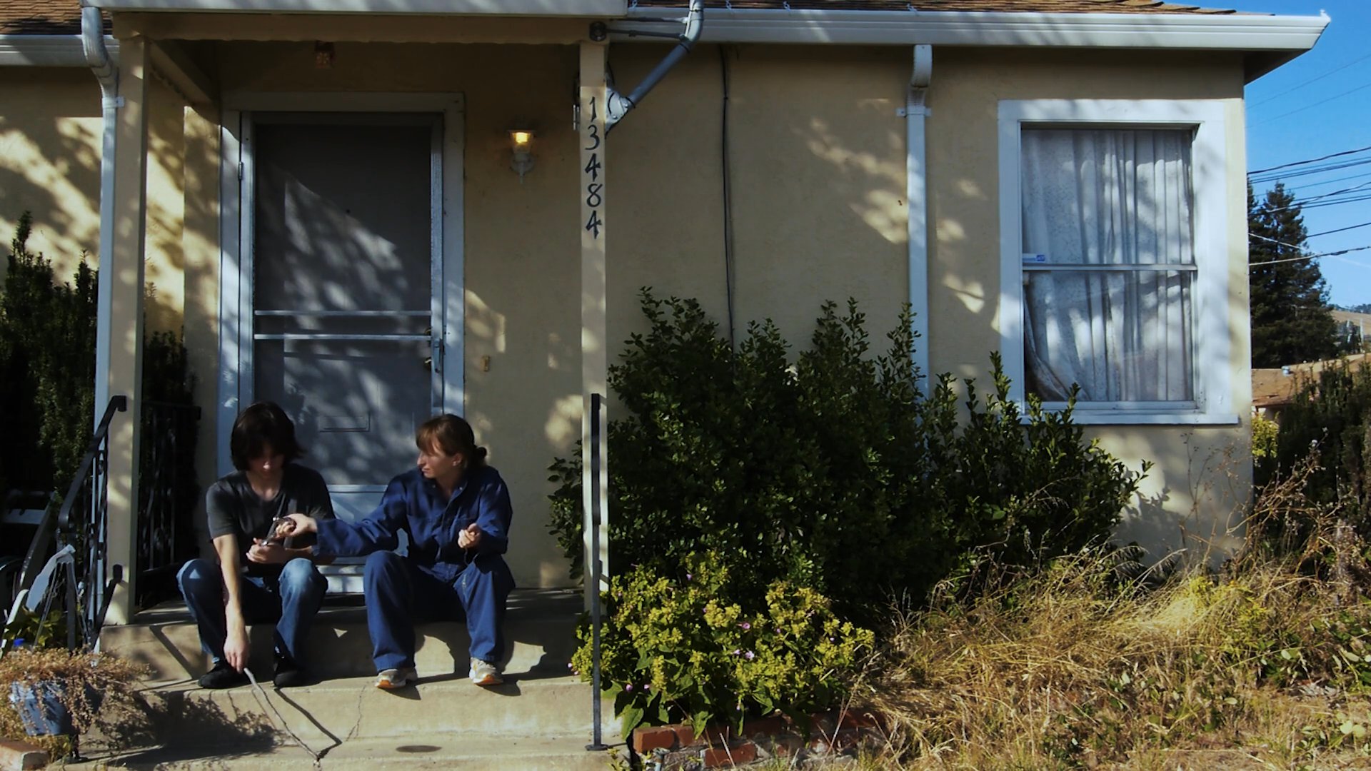 Two individuals discuss and perform diagnostic work on a vehicle engine while seated in front of a residential home.