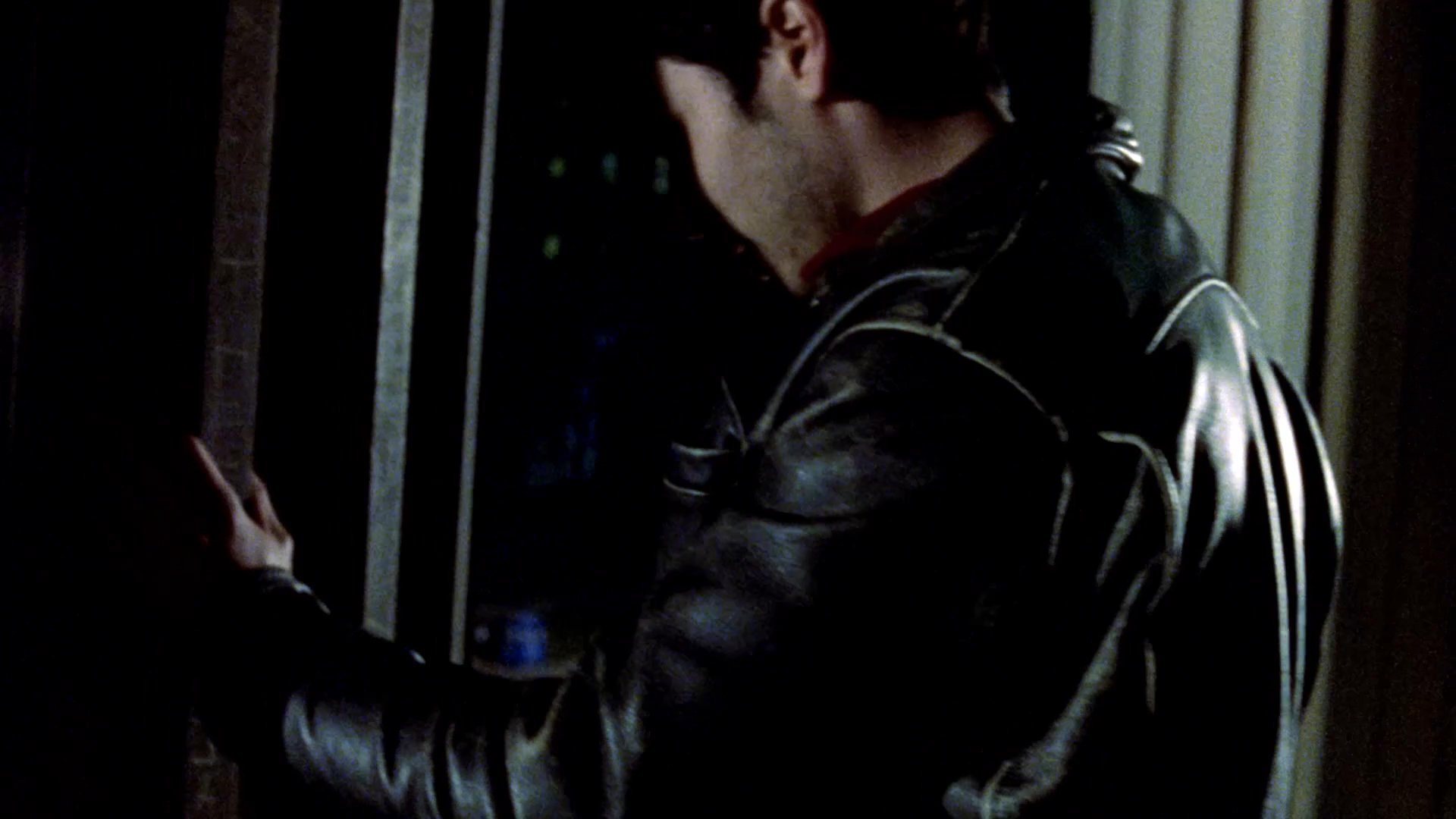 A man in a leather jacket gazes out over a night cityscape from a balcony, while a woman appears by the water's edge nearby. Their interactions suggest a contemplative and quiet atmosphere in an urban setting.
