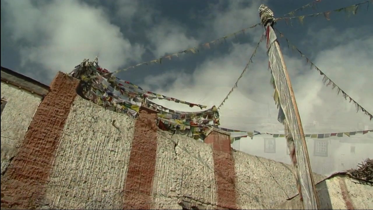 A panoramic view of a remote Himalayan village and monastery features traditional architecture, prayer flags, and rugged mountain terrain. The scene transitions to an interior courtyard where residents congregate near ancient stone structures.