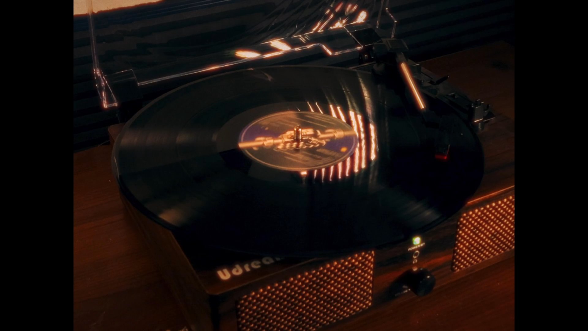 An individual sits at a computer desk in a dimly lit, warm-toned bedroom while a vinyl record rotates on a turntable nearby. The scene highlights a calm domestic environment with decorative shelf accents.