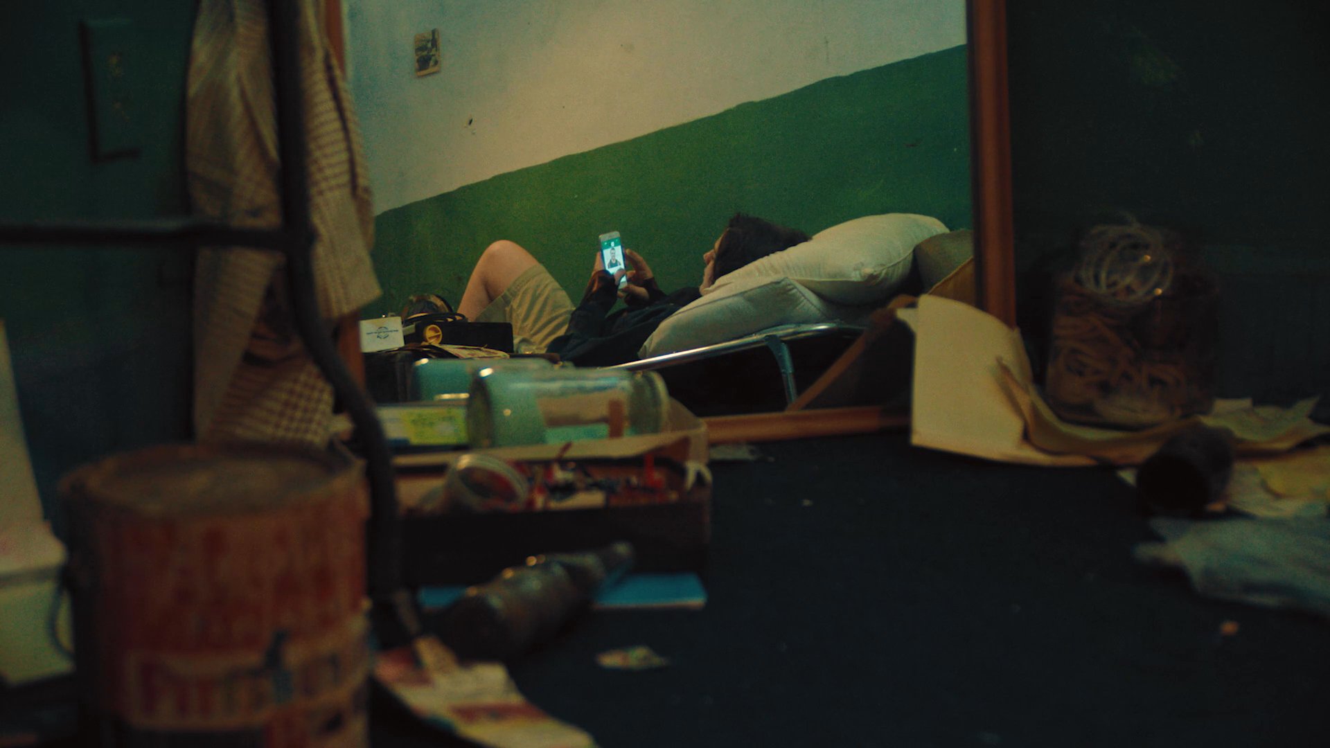 An individual reclines on a bed in a cluttered room, illuminated primarily by the glow of a smartphone screen. The scene emphasizes a quiet, secluded moment within a disheveled living environment.