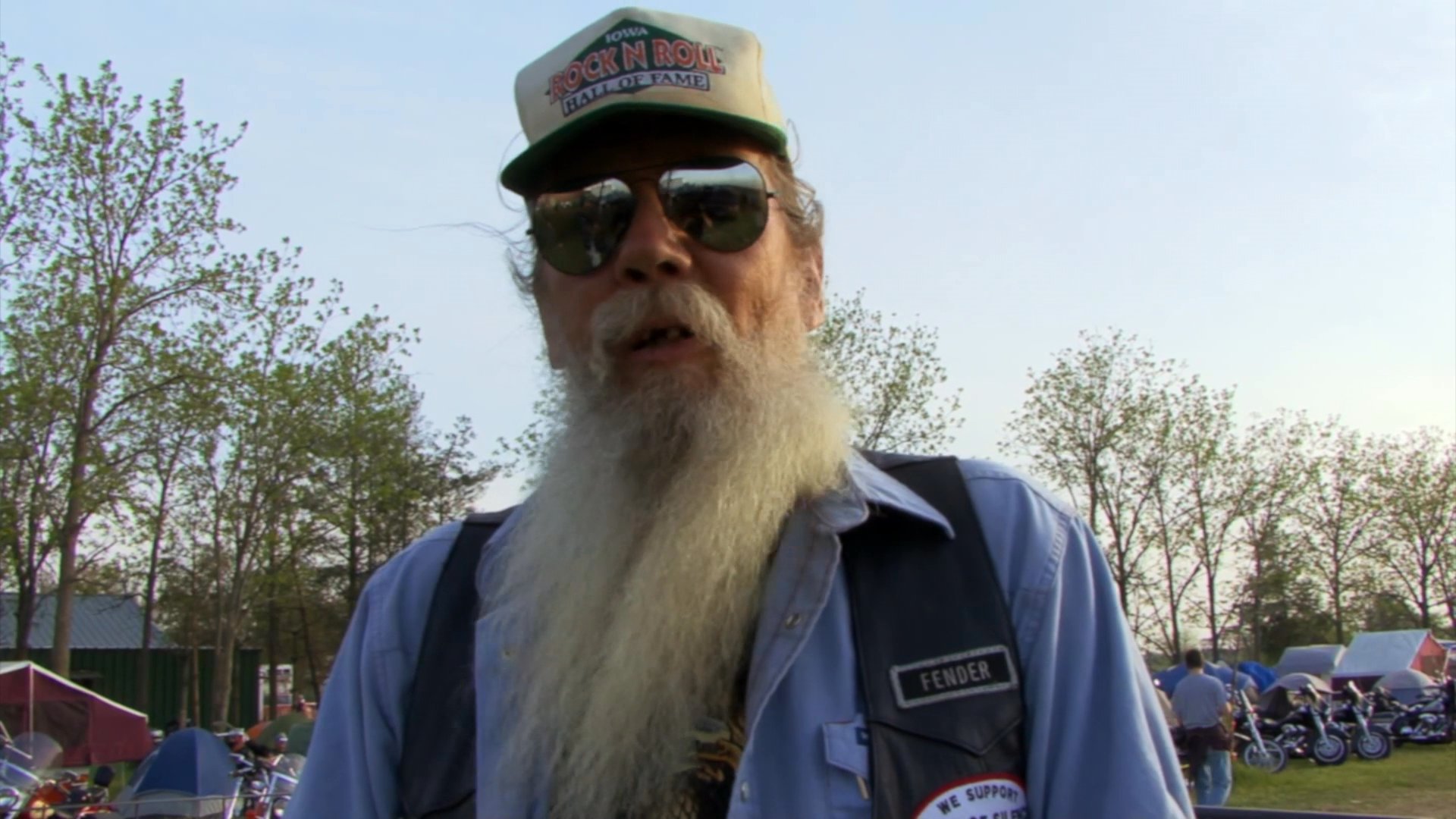 Attendees at a motorcycle rally share their experiences in an outdoor setting surrounded by fellow enthusiasts and parked bikes.