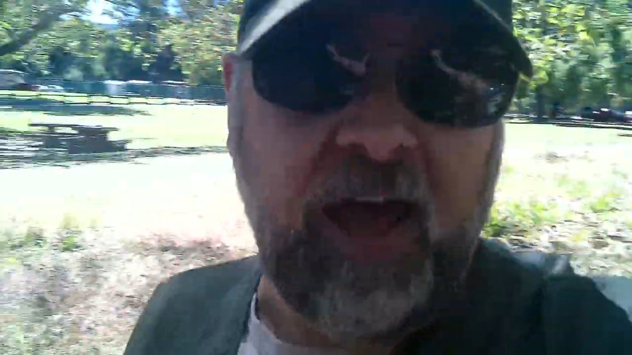 A man wearing sunglasses and a cap records a selfie-style vlog while walking through a wooded park area.