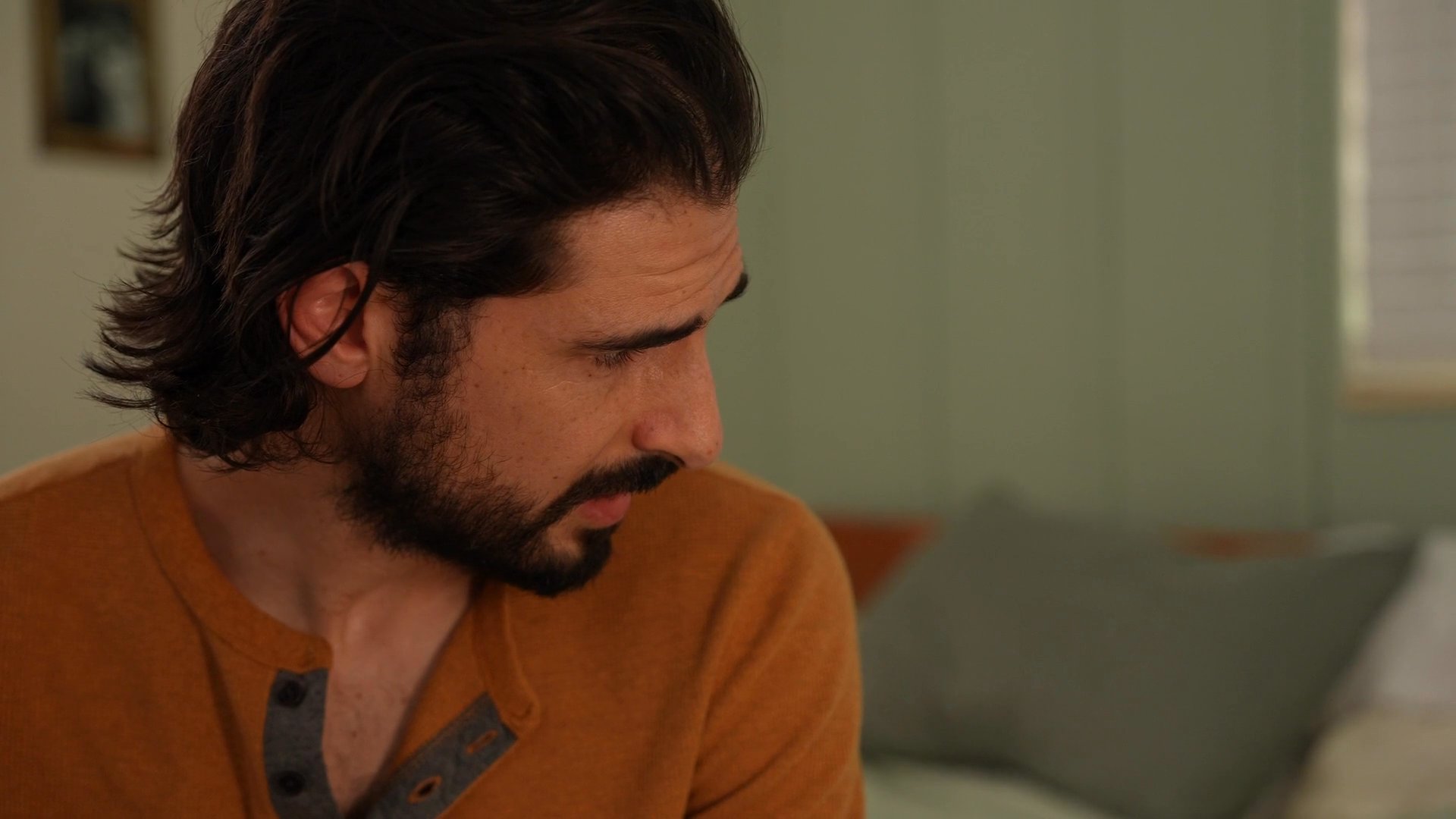 A man observes a woman resting in bed, establishing a moment of shared quiet and emotional intimacy. The scene captures a soft, personal interaction within a domestic bedroom setting.