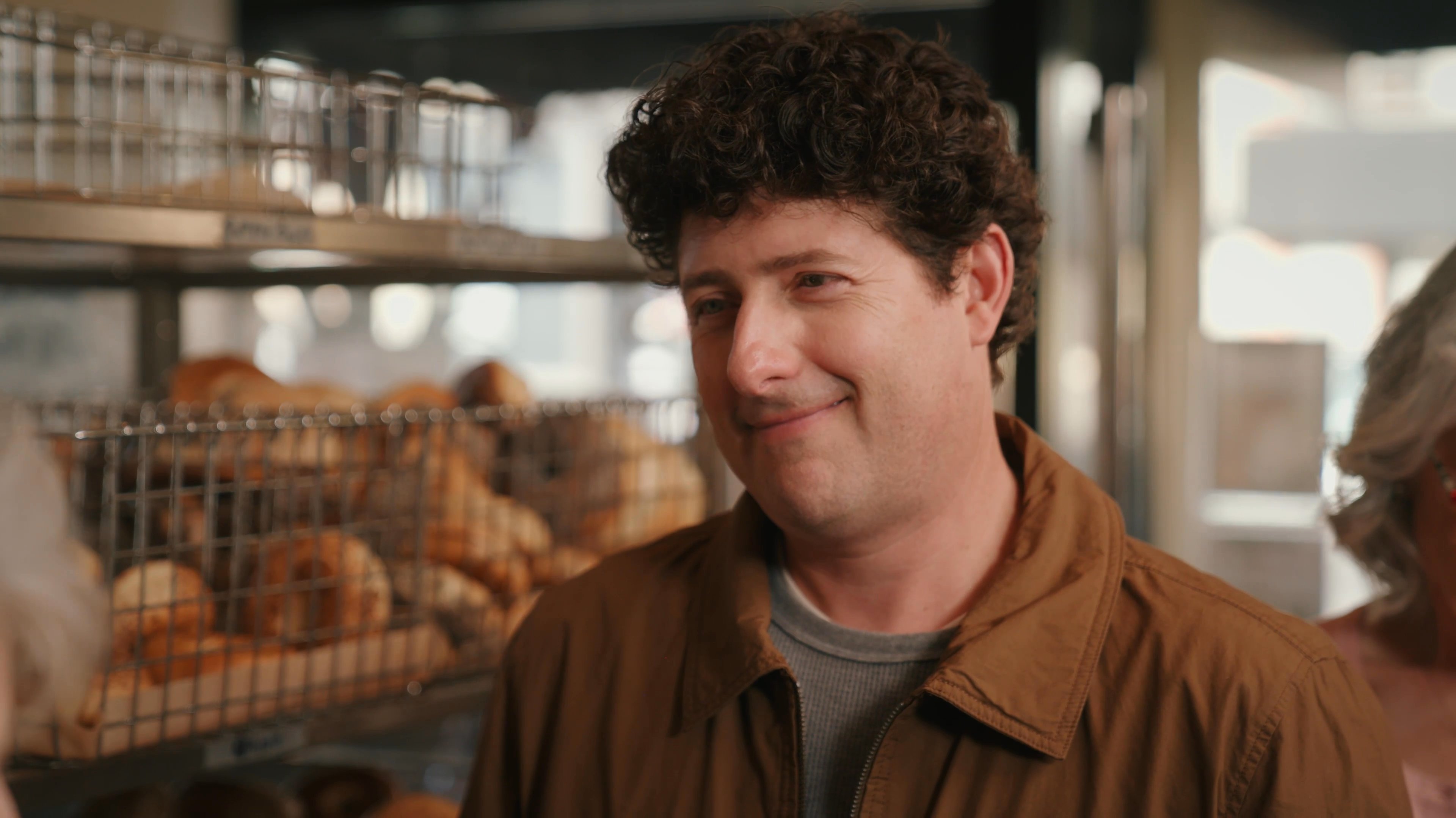 An elderly woman engages in an animated conversation with a man inside a busy bakery. The pair discusses options while standing in front of shelves stocked with baked goods.