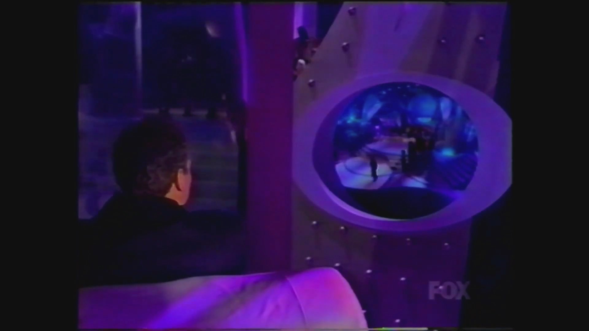 A formal awards show host presents content on stage while interacting with an off-screen partner through a circular monitor display.
