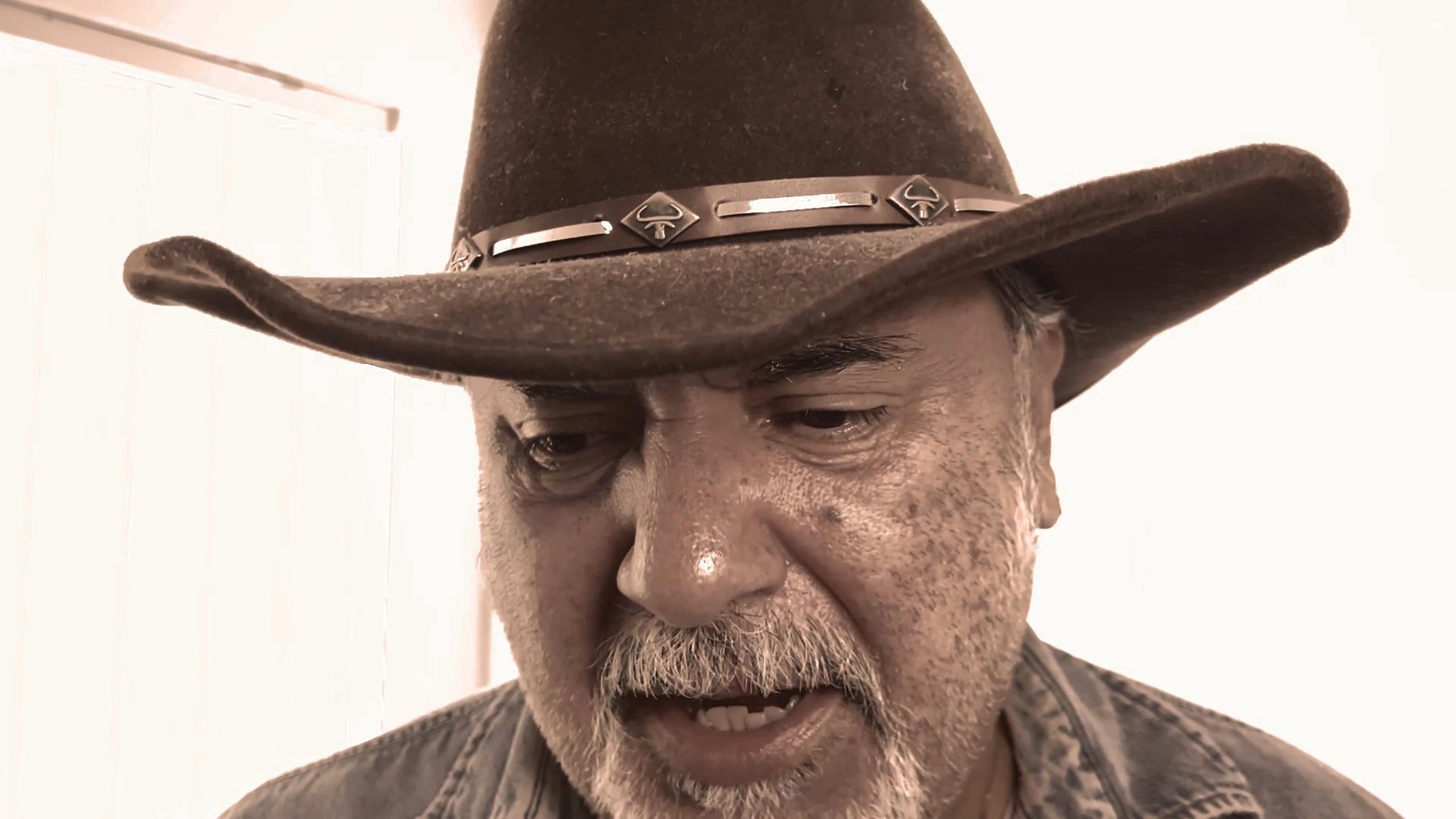 A man wearing a cowboy hat delivers a focused monologue while looking directly into the camera. He utilizes varying facial expressions to convey his message throughout the performance.
