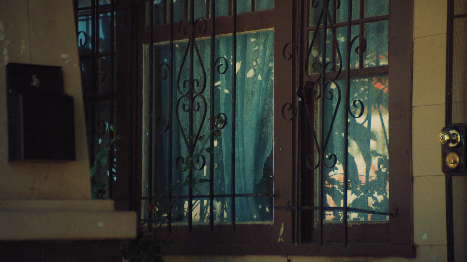 Two individuals engage in a conversation while sitting inside a vehicle parked on a residential street. The focus remains on their interaction as they sit before a house with a gated window.