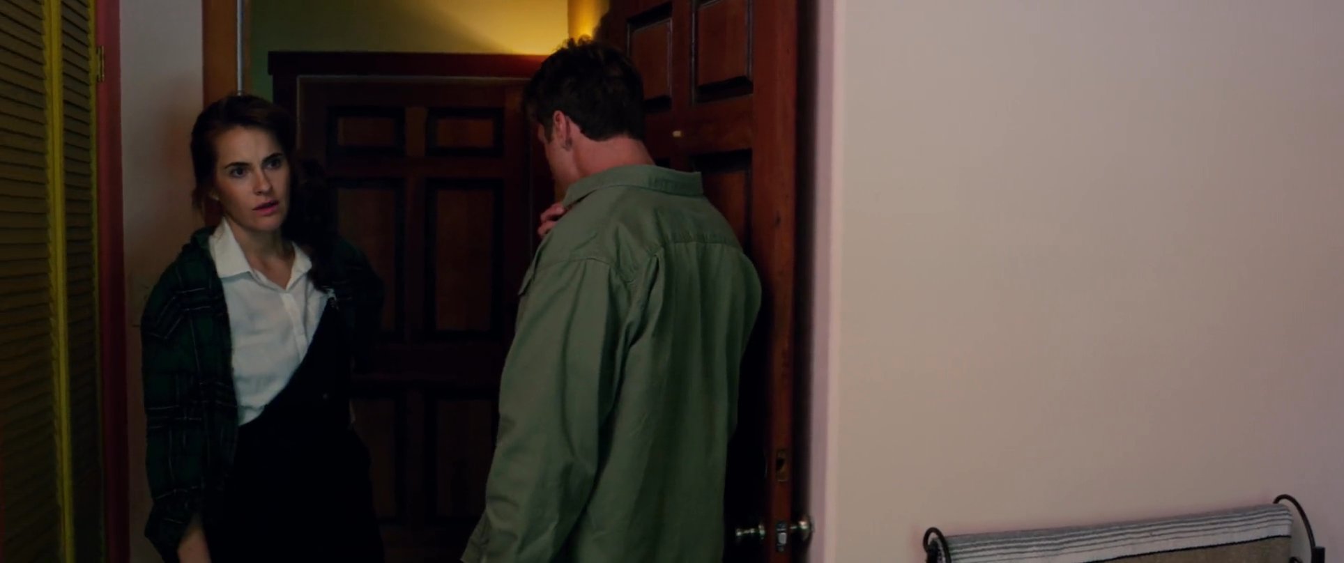 A man and a woman engage in a serious, quiet conversation within a room while a dishevelled individual sits nearby. The interaction appears charged with underlying tension and unspoken conflict.