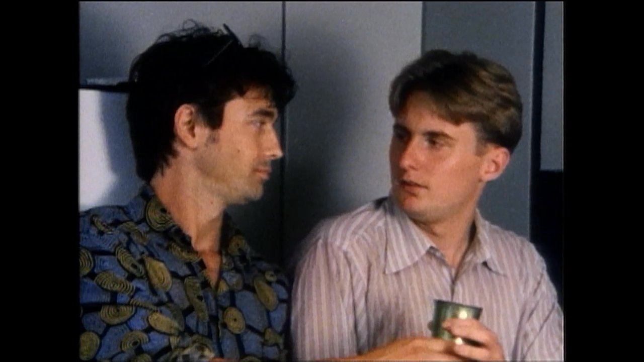Two men engage in a conversation while standing in a kitchen setting, with one offering a beverage to the other. They interact naturally as they exchange words in a casual indoor environment.