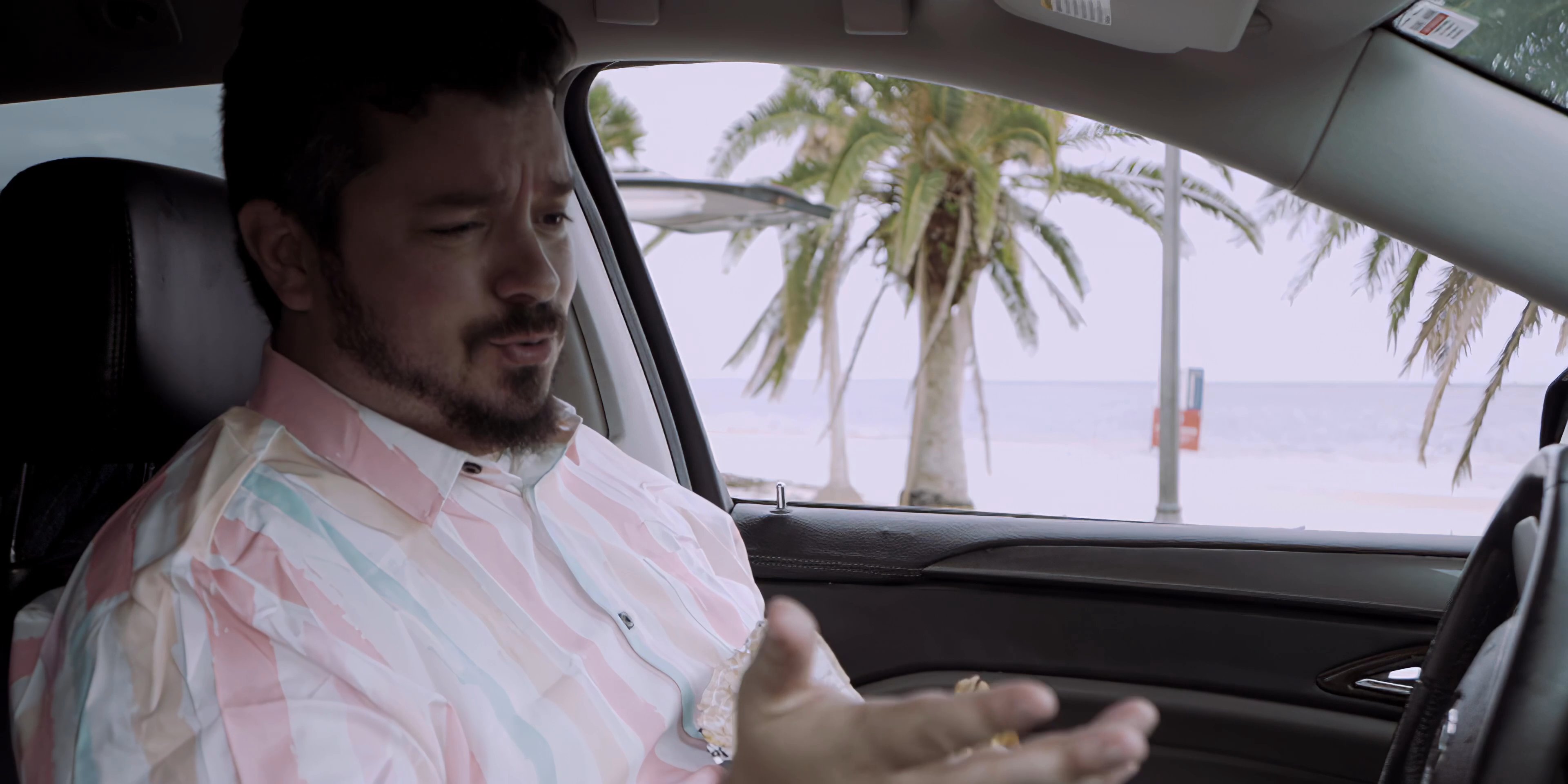 A man in his car engages in a remote video conversation while parked near a tropical beach. He listens attentively to the speaker on his smartphone mounted to the dashboard while eating a meal.