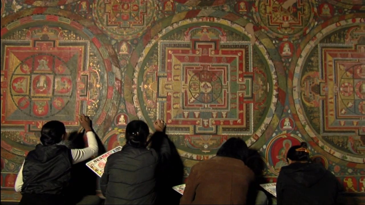 Art conservators meticulously clean and repair intricate ancient murals inside a Buddhist monastery. The process involves careful manual restoration of detailed religious iconography and geometric mandalas.