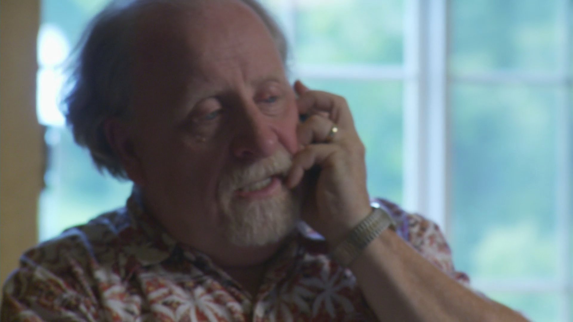 An elderly man appears visibly distressed while engaged in a telephone conversation at his desk. Meanwhile, a woman continues to work on her computer while also participating in a phone call nearby.