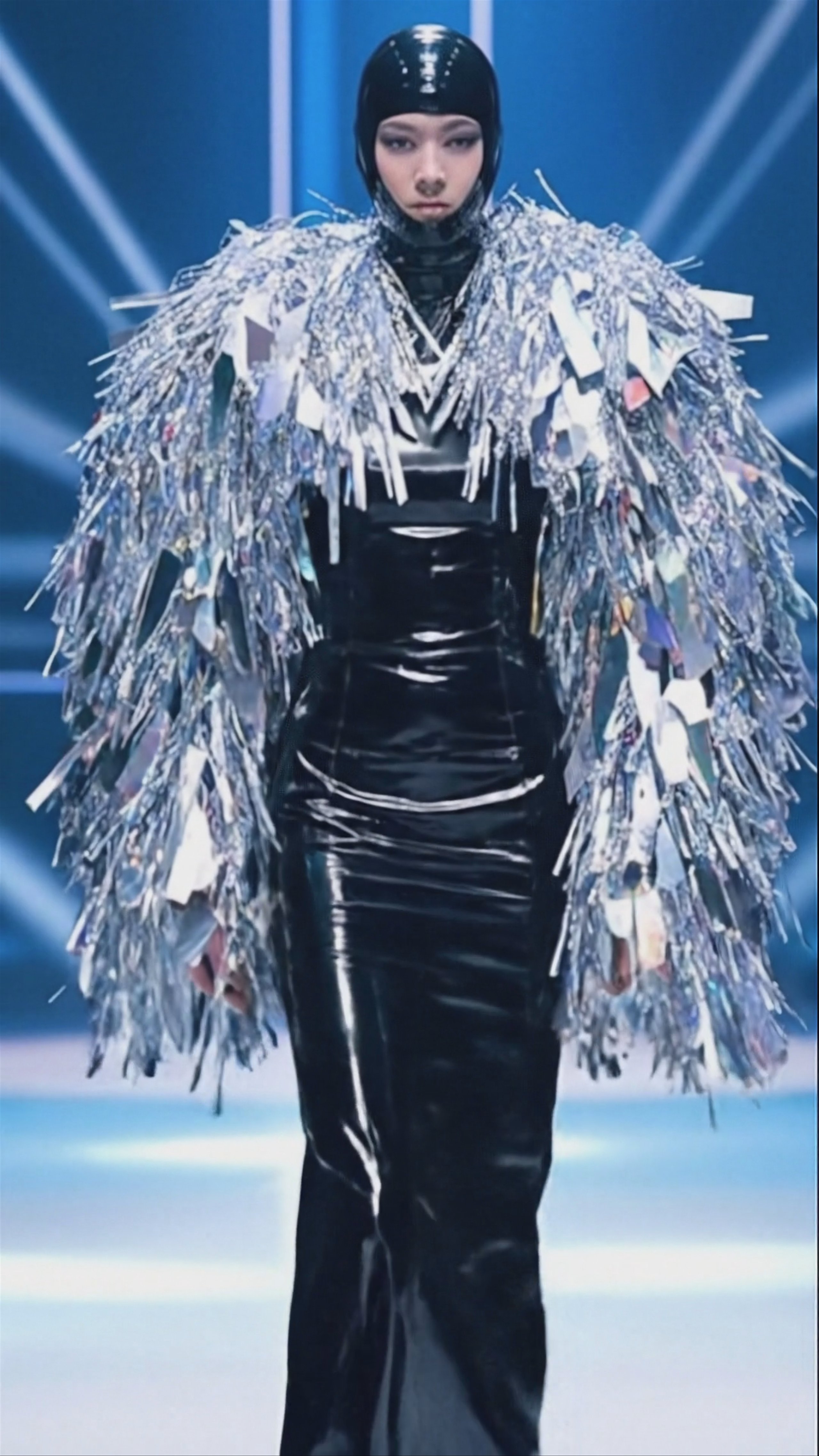A fashion model stands on a runway wearing a glossy black latex hood and gown paired with a massive, textured silver tinsel jacket. The background features cool blue stage lighting and bright vertical beams. Generated using Veo 3.1 Fast at a vertical 1080x1920 resolution.