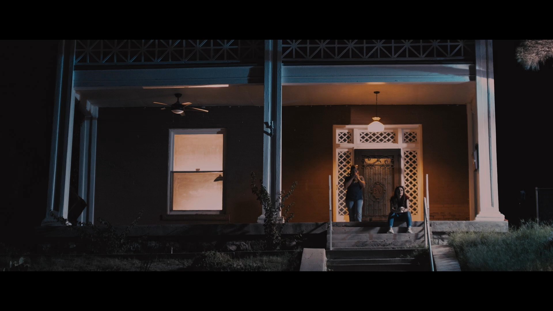 Two women sit together on a residential porch at night, sharing a quiet moment and a warm drink. One woman hands a mug to the other while they speak softly about a sleeping family member.