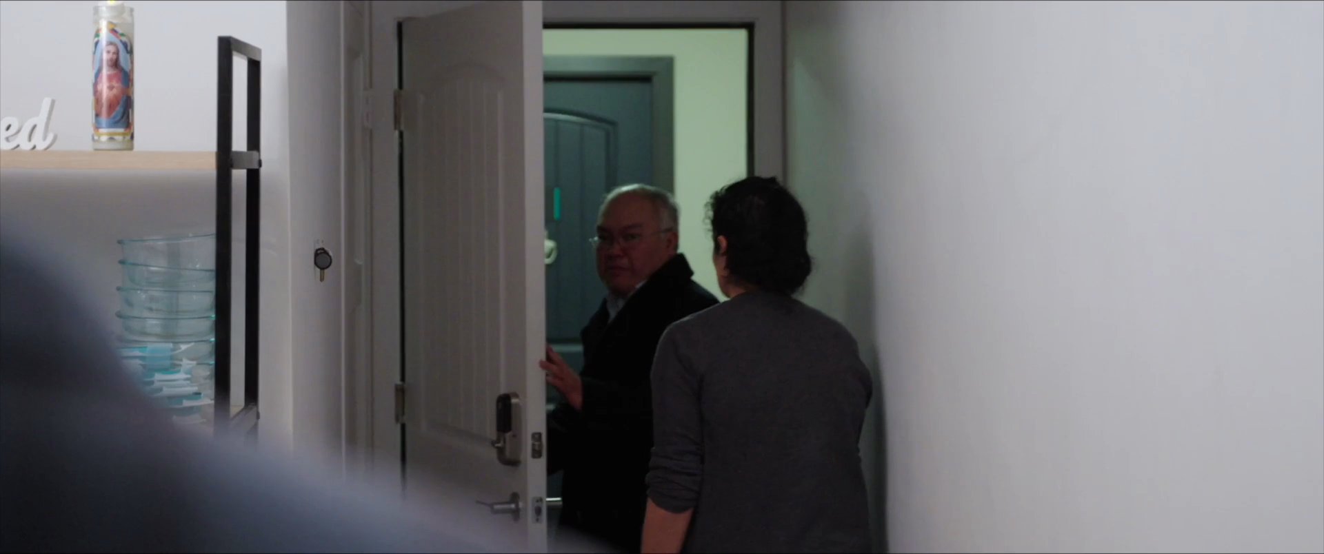 A young man departs from a home, leaving his family behind as he contemplates his situation while sitting in a car. The sequence captures a somber moment of transition marked by lingering glances and emotional distance.