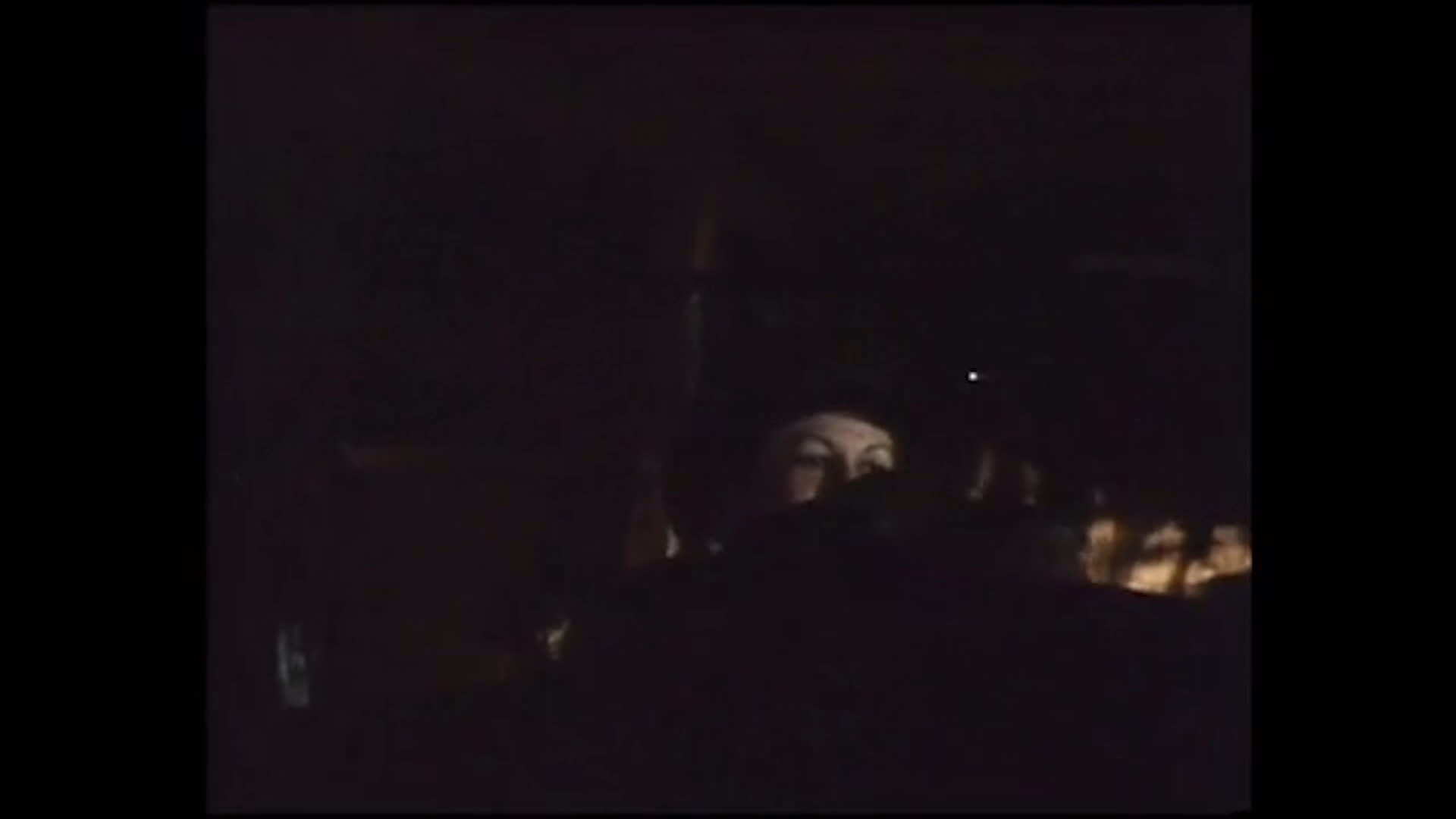 A stable worker in a dark blue jacket walks through a dim barn and tends to a horse, handling its tack near the stall. The worker describes 15 years on the job and the importance of having room and horse boarding included in the arrangement.