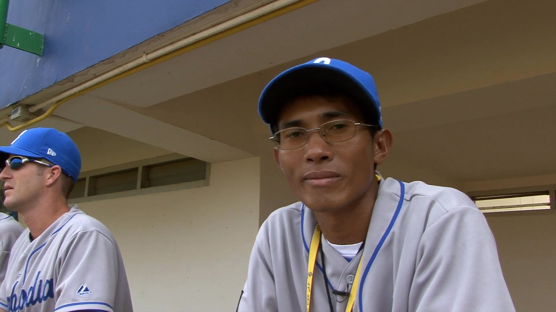 A baseball player wearing a team uniform and cap discusses a game between the Philippines and Cambodia while seated in a dugout.