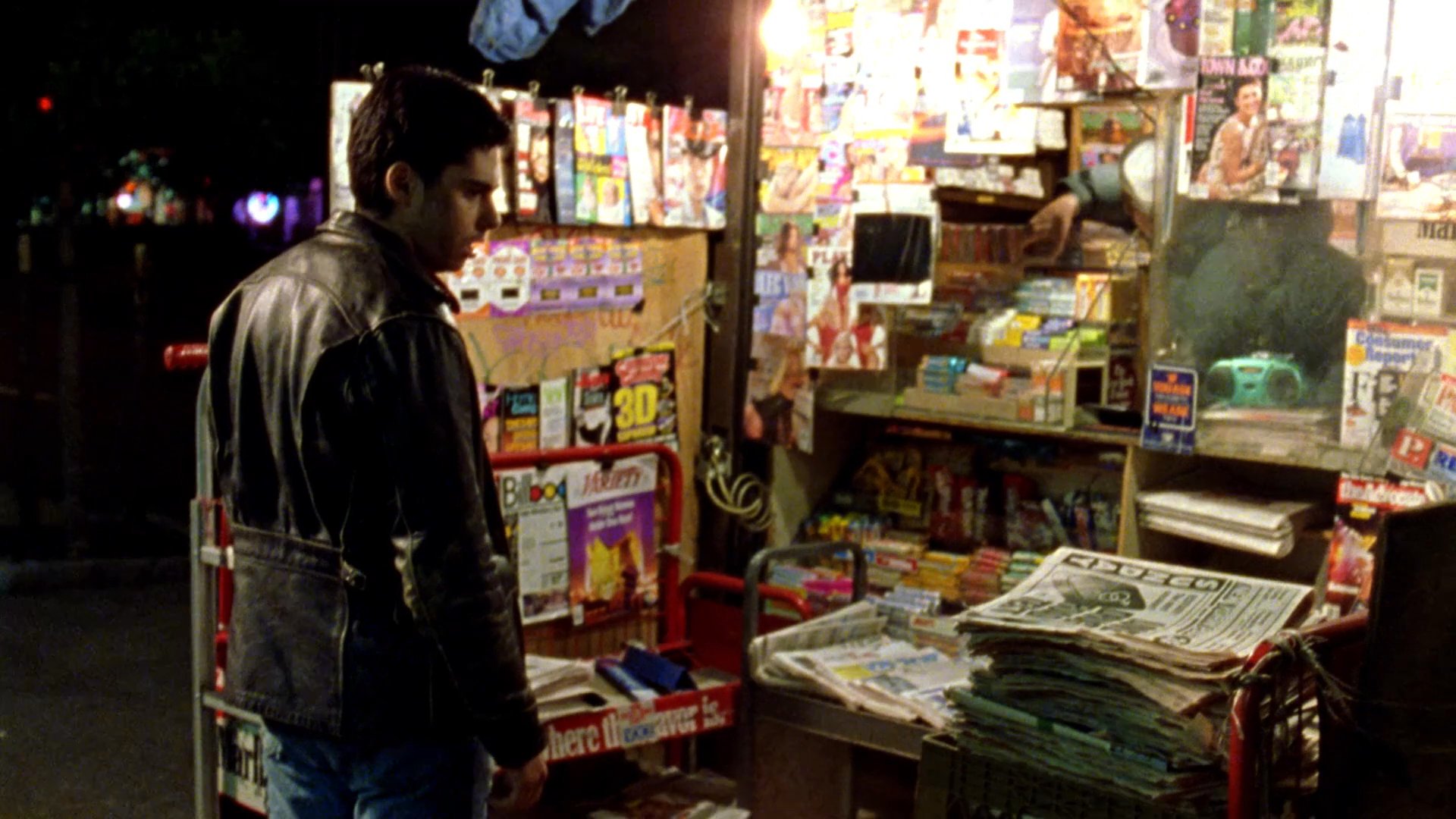 A man approaches an urban newsstand at night to examine and read a local newspaper. The scene highlights the contrast between the dimly lit city surroundings and the brightly displayed printed media.
