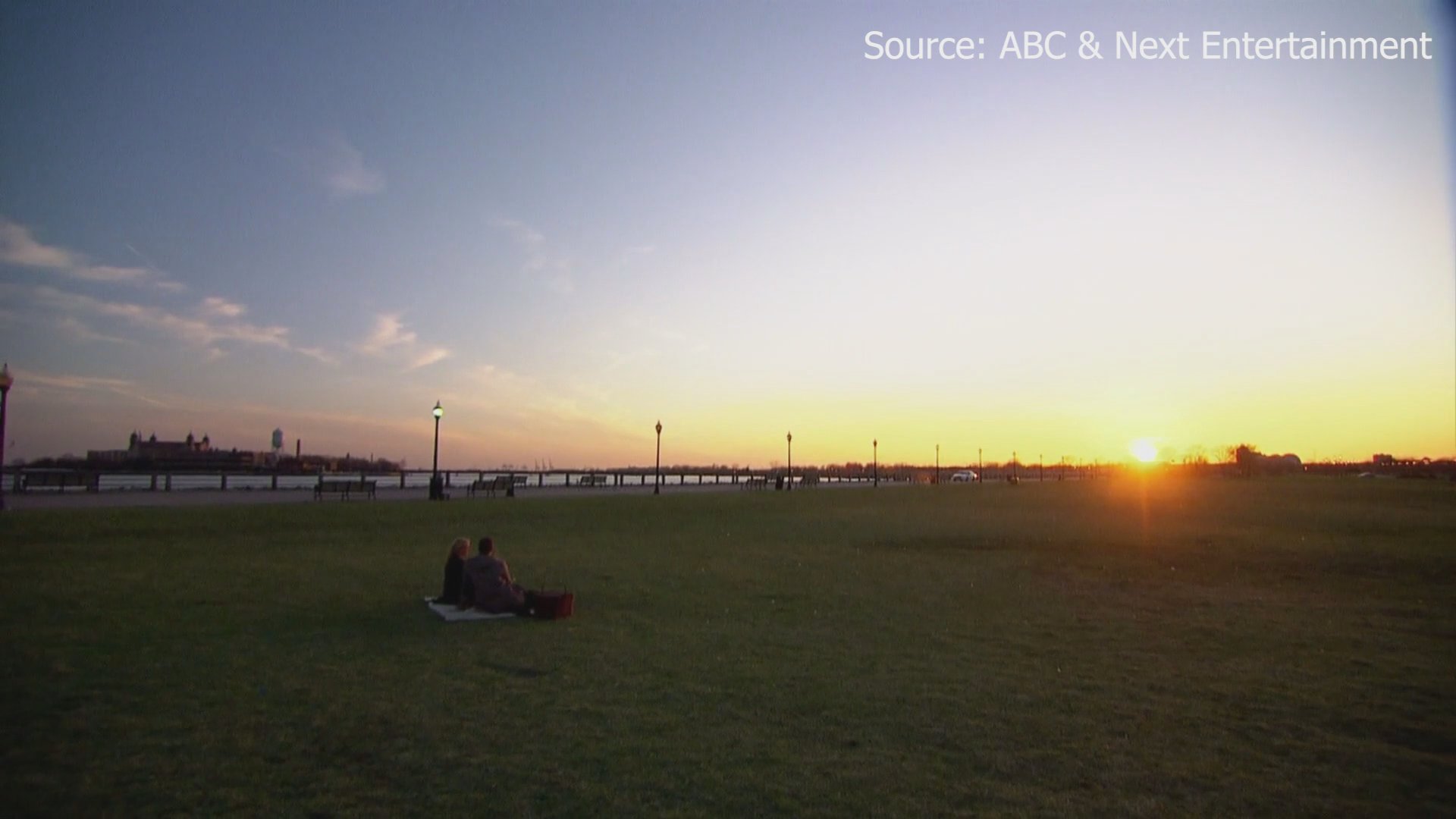 A couple enjoys a private champagne picnic in a waterfront park with a helicopter nearby and the New York City skyline at sunset in the background.