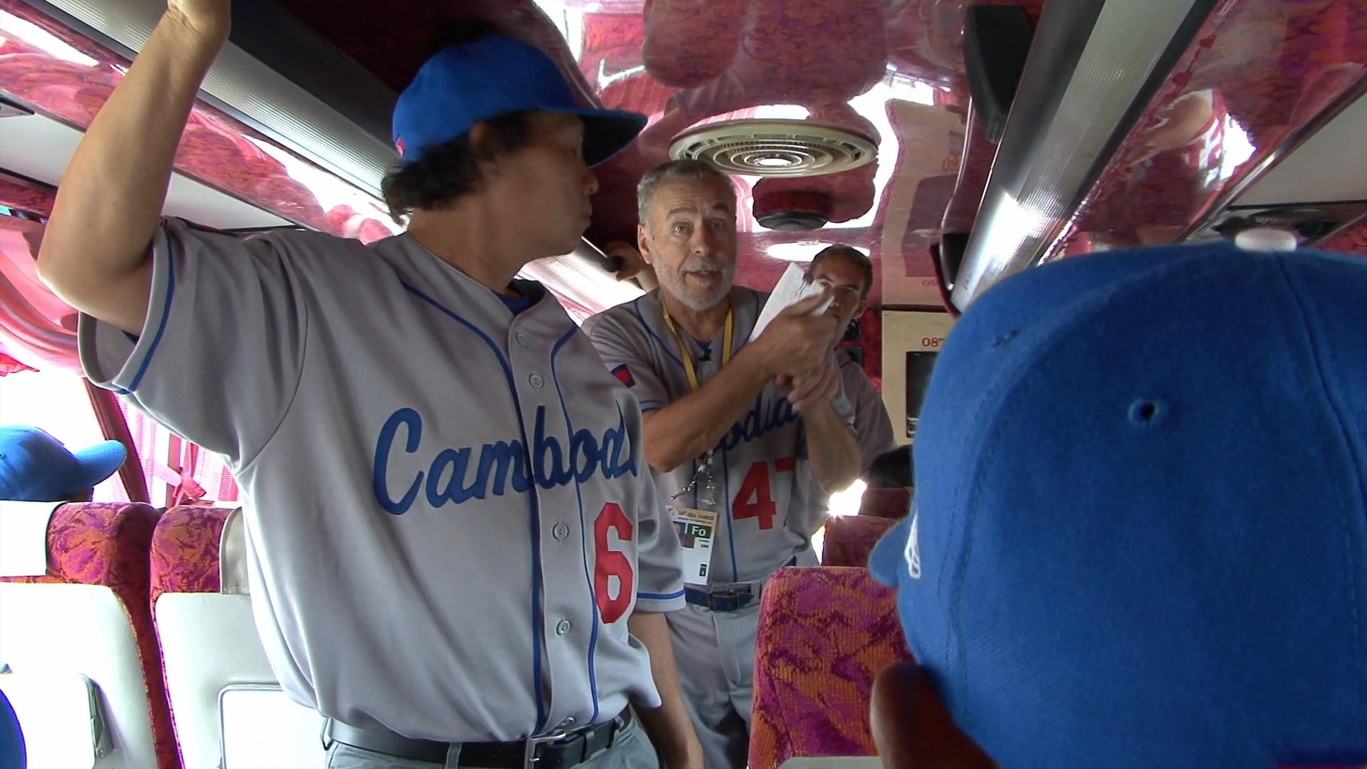 A baseball player wearing a team jersey demonstrates a batting stance while standing in the aisle of a bus, engaging his teammates during a group journey.